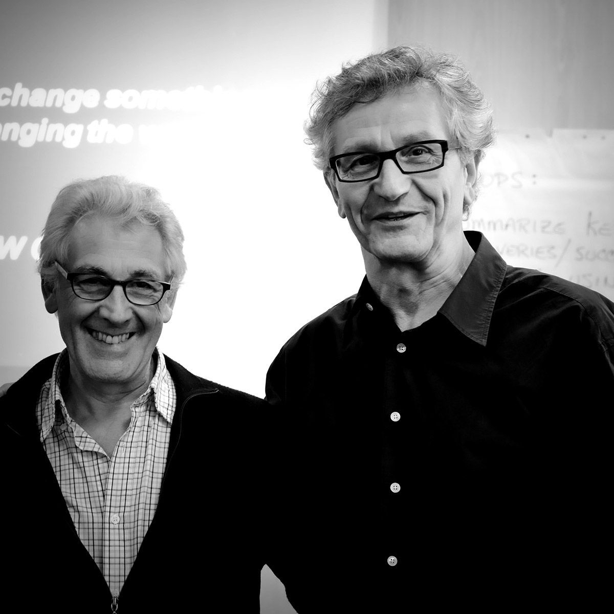 Deux hommes souriants, portant des lunettes, devant un tableau blanc. L'un est en chemise boutonnée, l'autre en chemise sombre.