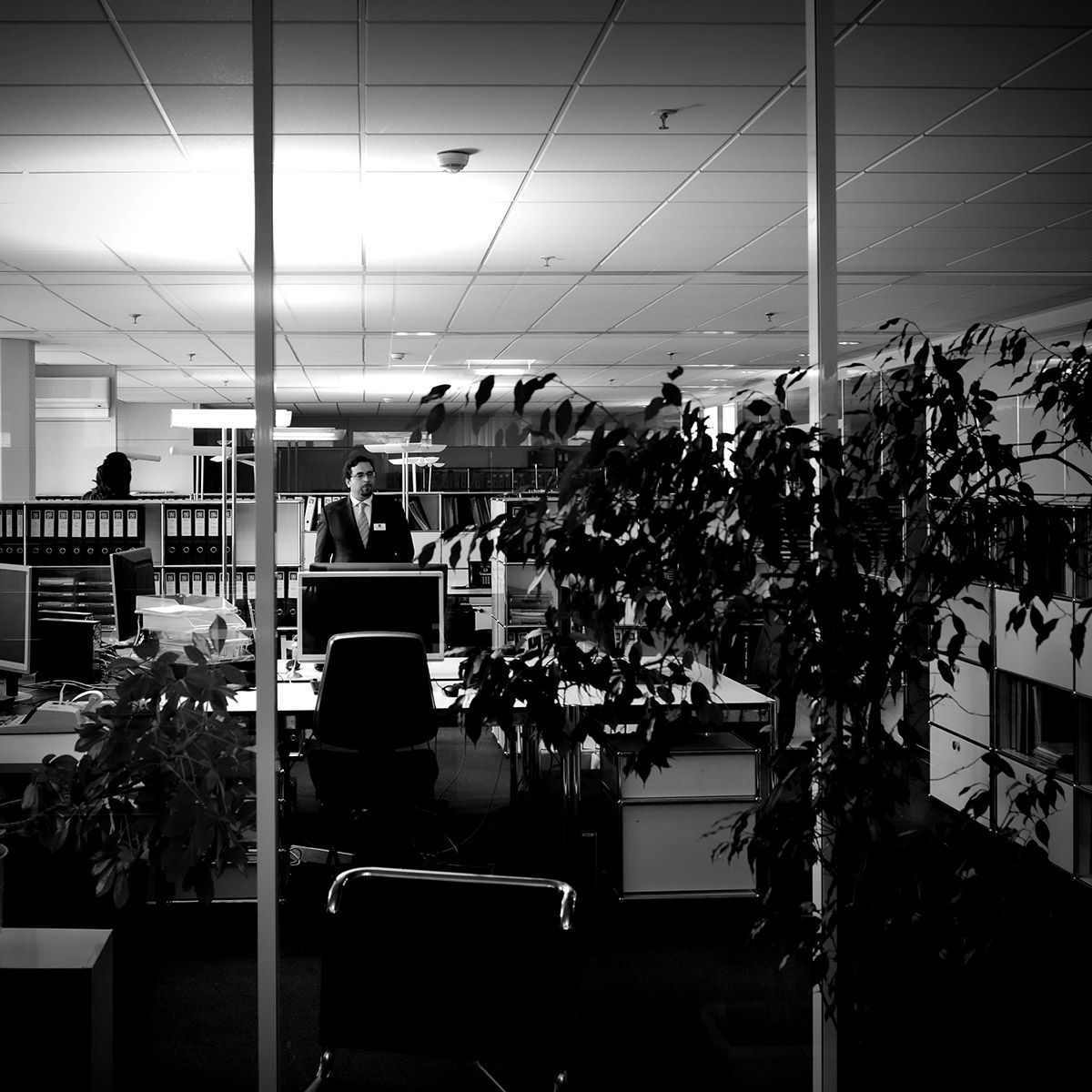 Intérieur de bureau avec cloisons, plantes et une personne debout derrière un bureau. Noir et blanc.