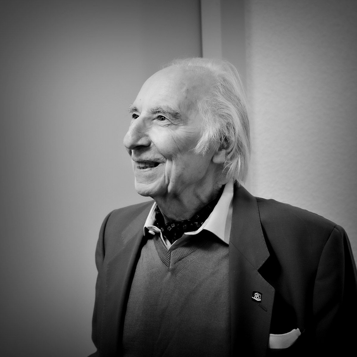 Un homme âgé souriant, vêtu d'un blazer, d'un pull et d'une écharpe. Fond clair.