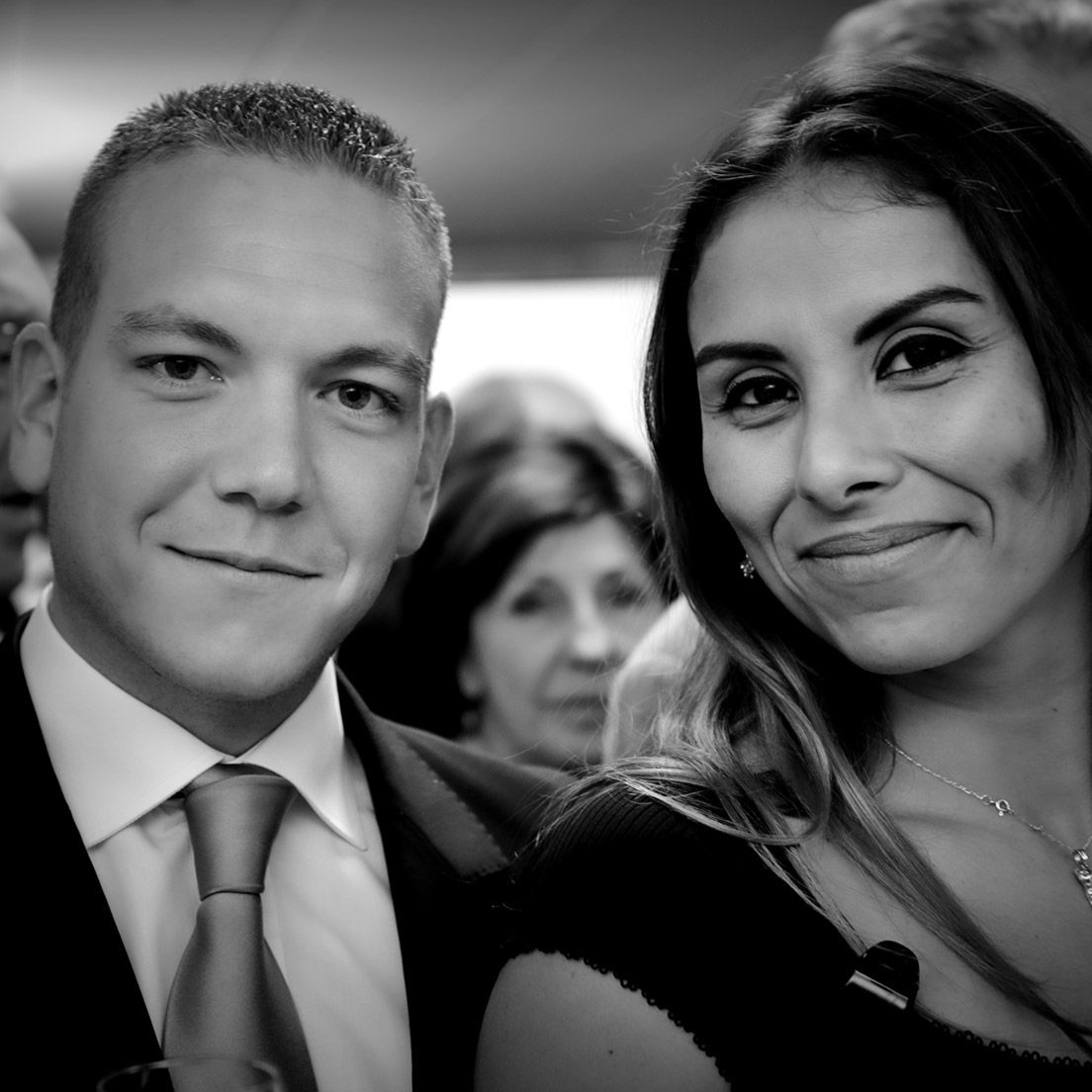 Un homme et une femme sourient, posant pour une photo. Ils se trouvent dans une pièce bondée, avec d'autres personnes en arrière-plan.