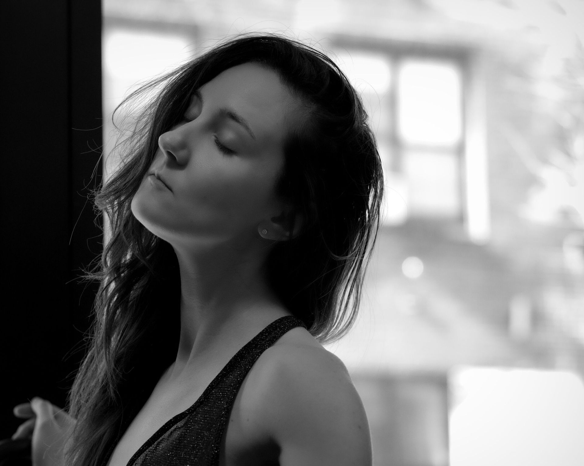 Femme aux yeux fermés, la tête inclinée vers une fenêtre, lumière du soleil, vêtue d'un haut sombre, photo en noir et blanc.