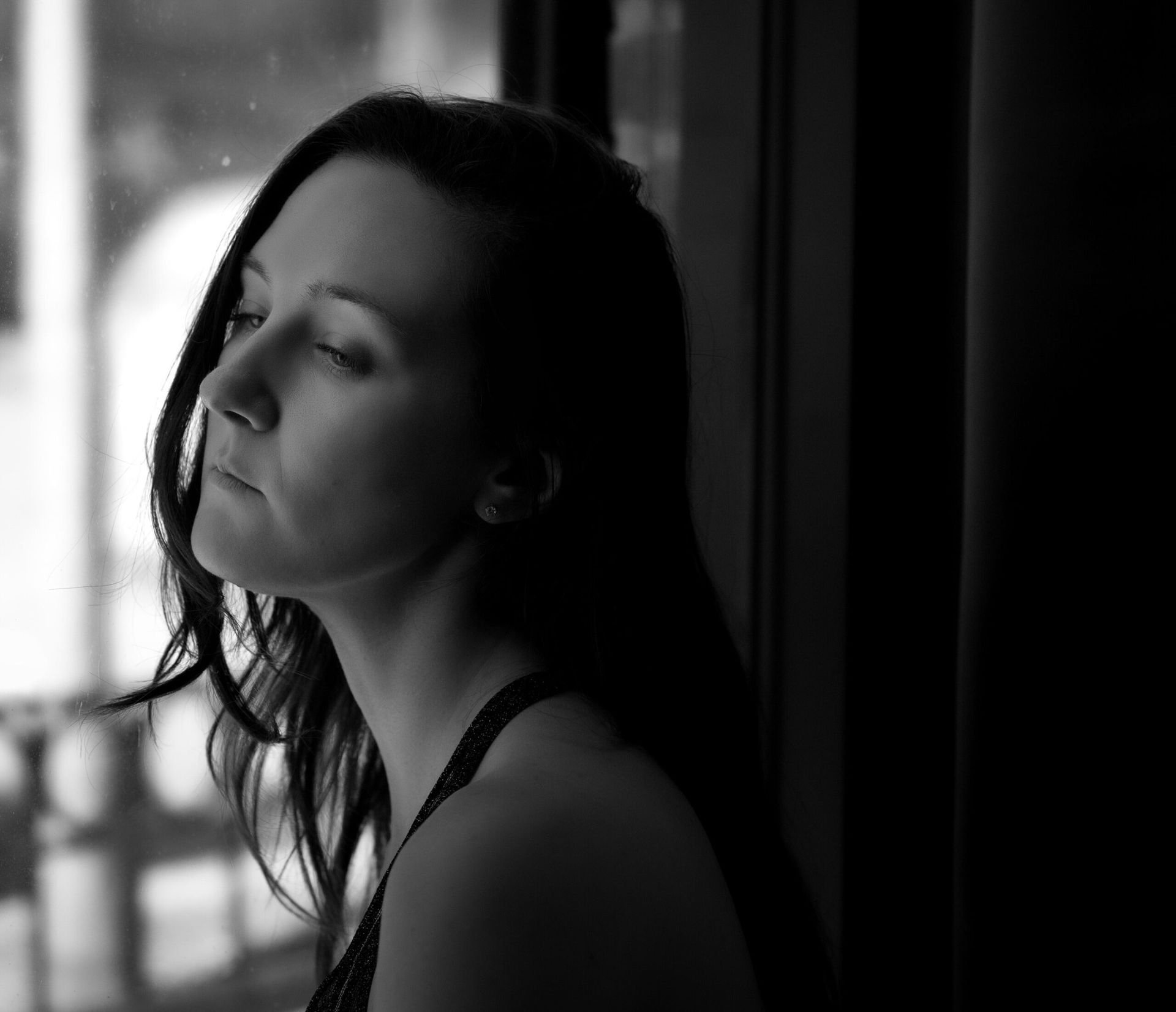 Femme aux longs cheveux noirs, les yeux fermés, regardant par la fenêtre, en noir et blanc.
