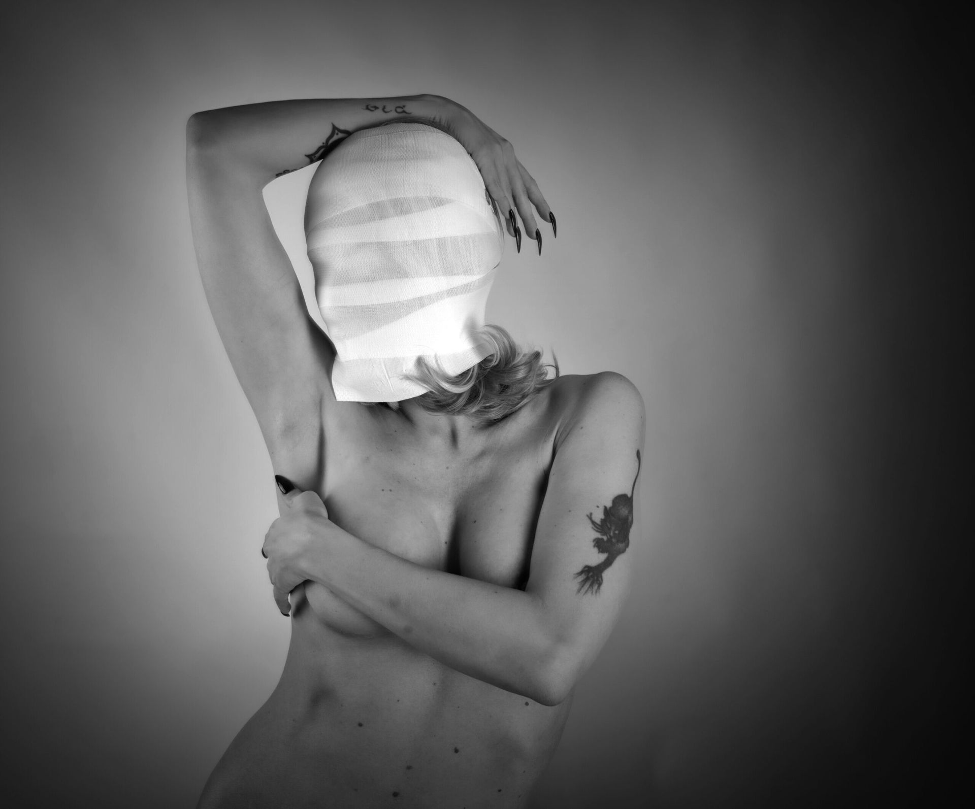 Femme au visage bandé, nue jusqu'à la taille, les bras levés et tatoués.
