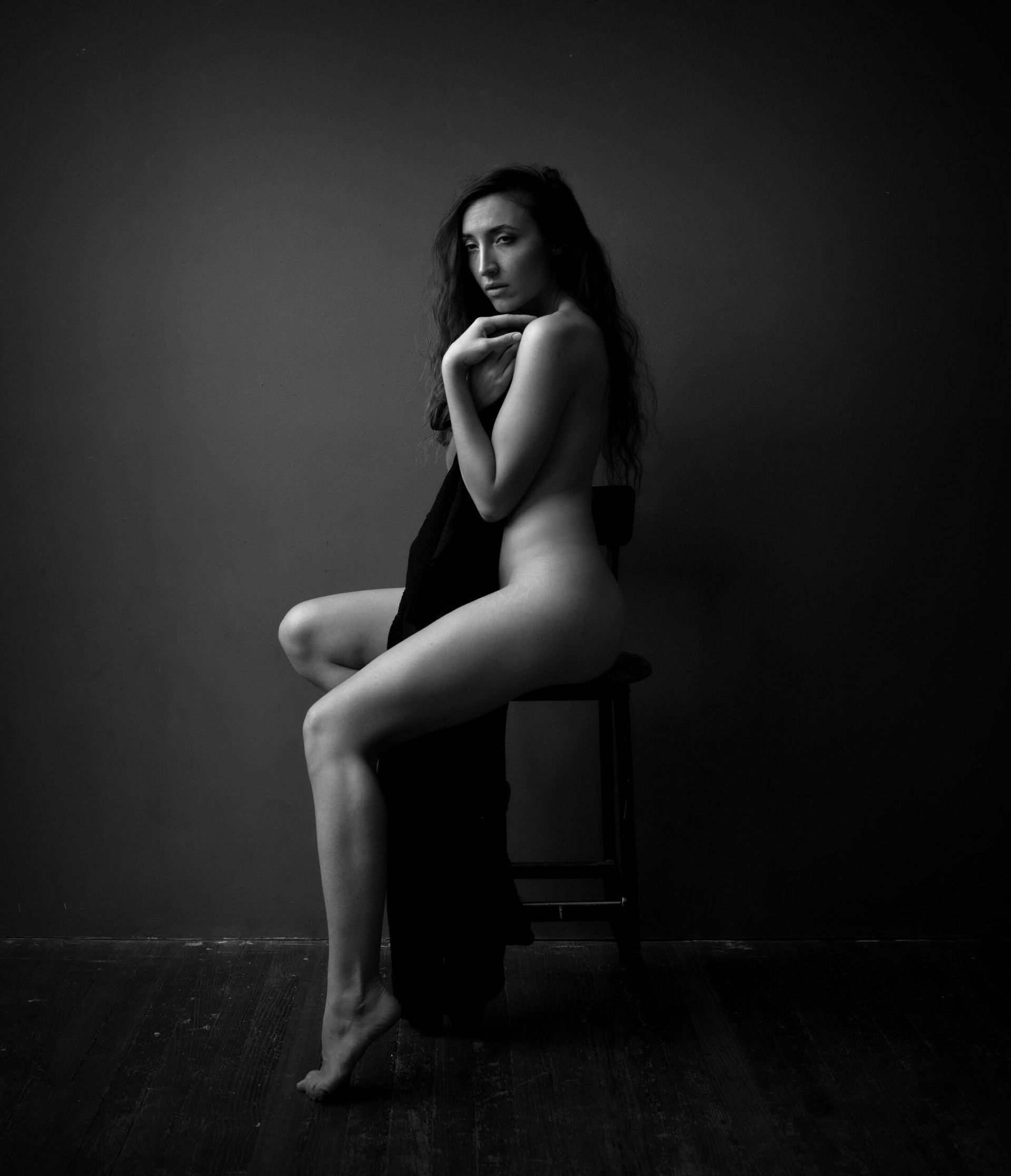 Une personne nue est assise sur un tabouret, recouverte d'un drap sombre, éclairée dans une lumière dramatique en noir et blanc.