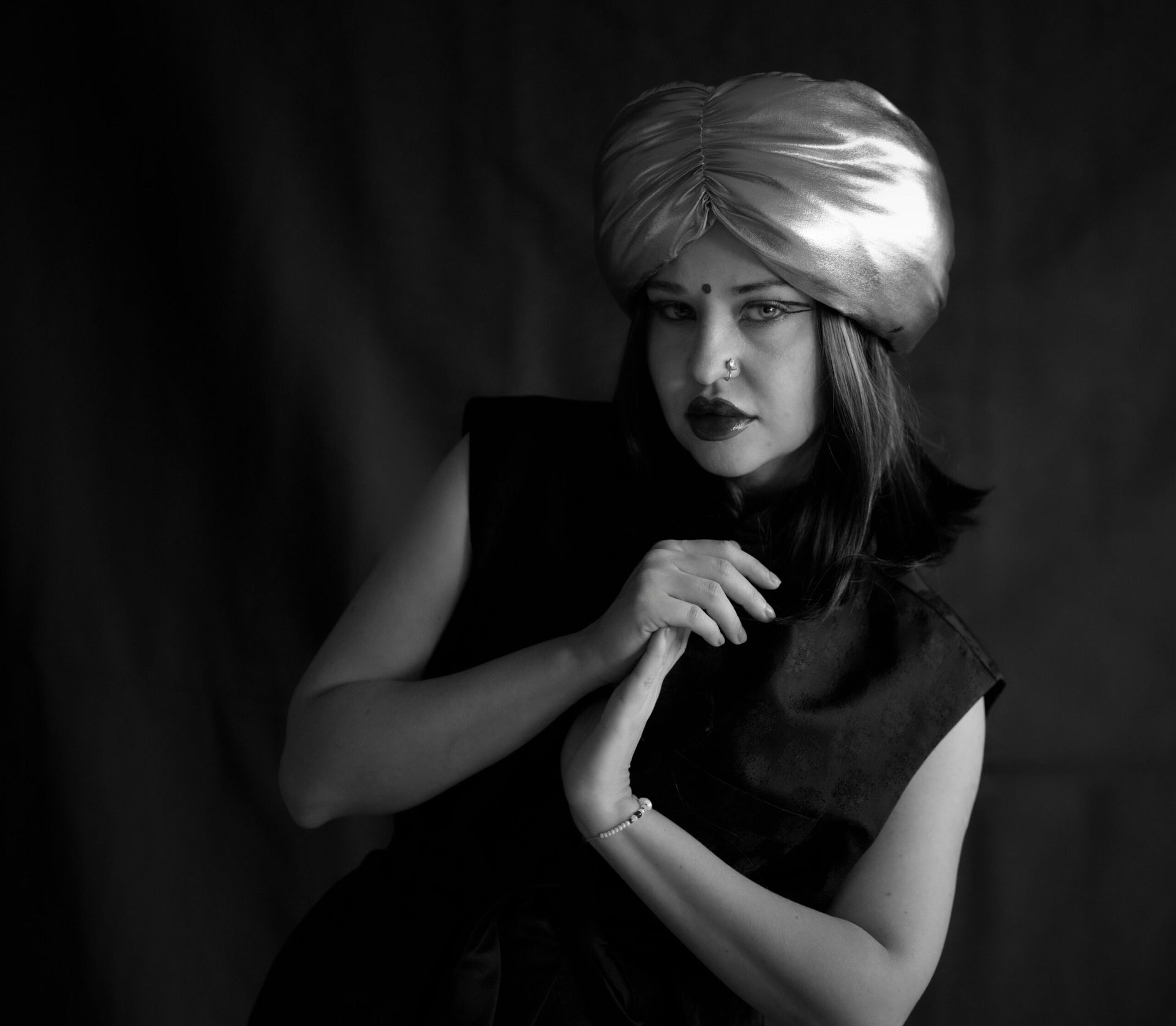 Femme portant un turban doré, robe sombre, mains jointes, décor sombre, expression intense.