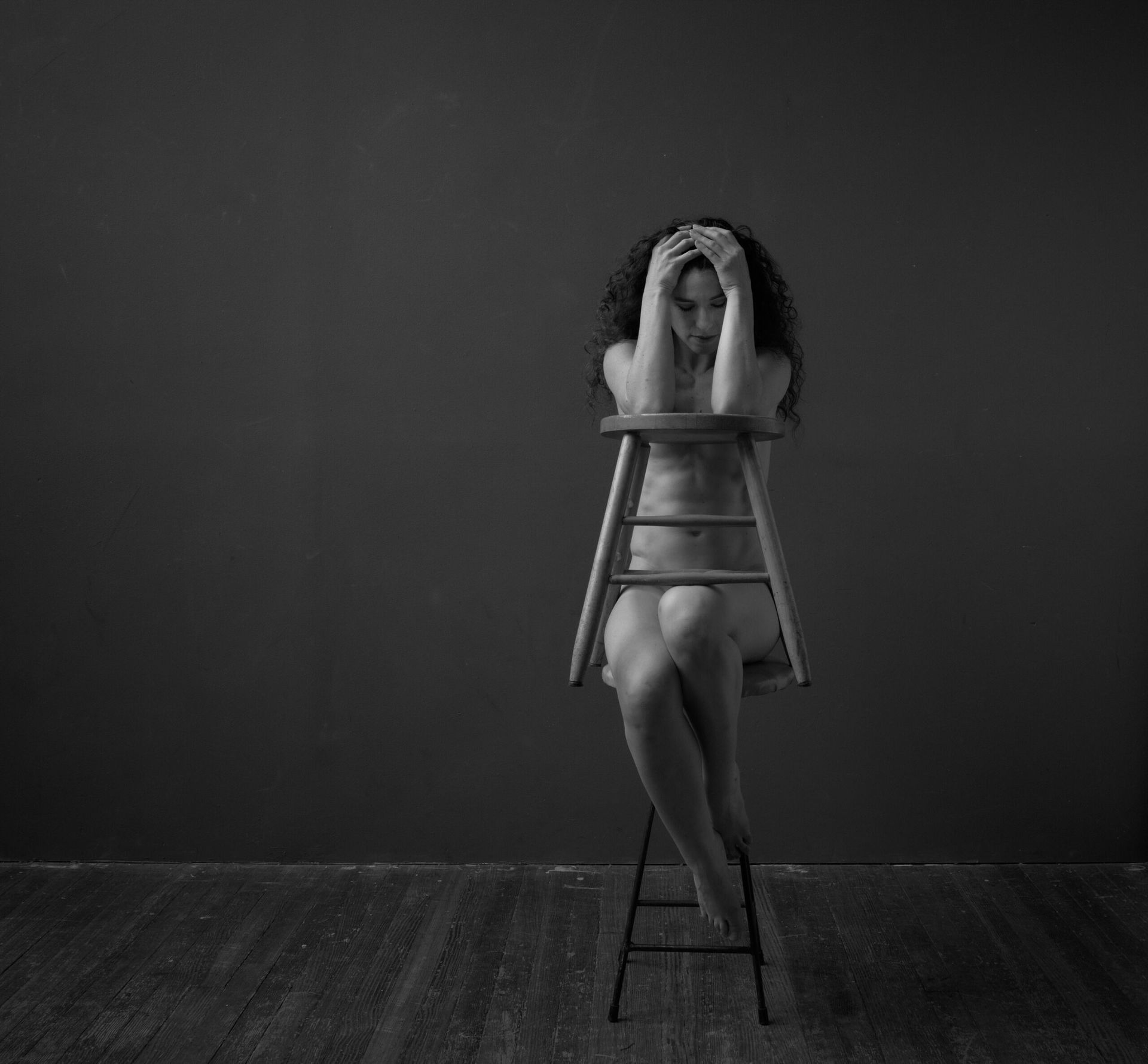 Une personne nue assise sur un tabouret, les mains sur la tête, le regard baissé, dans une pièce grise.