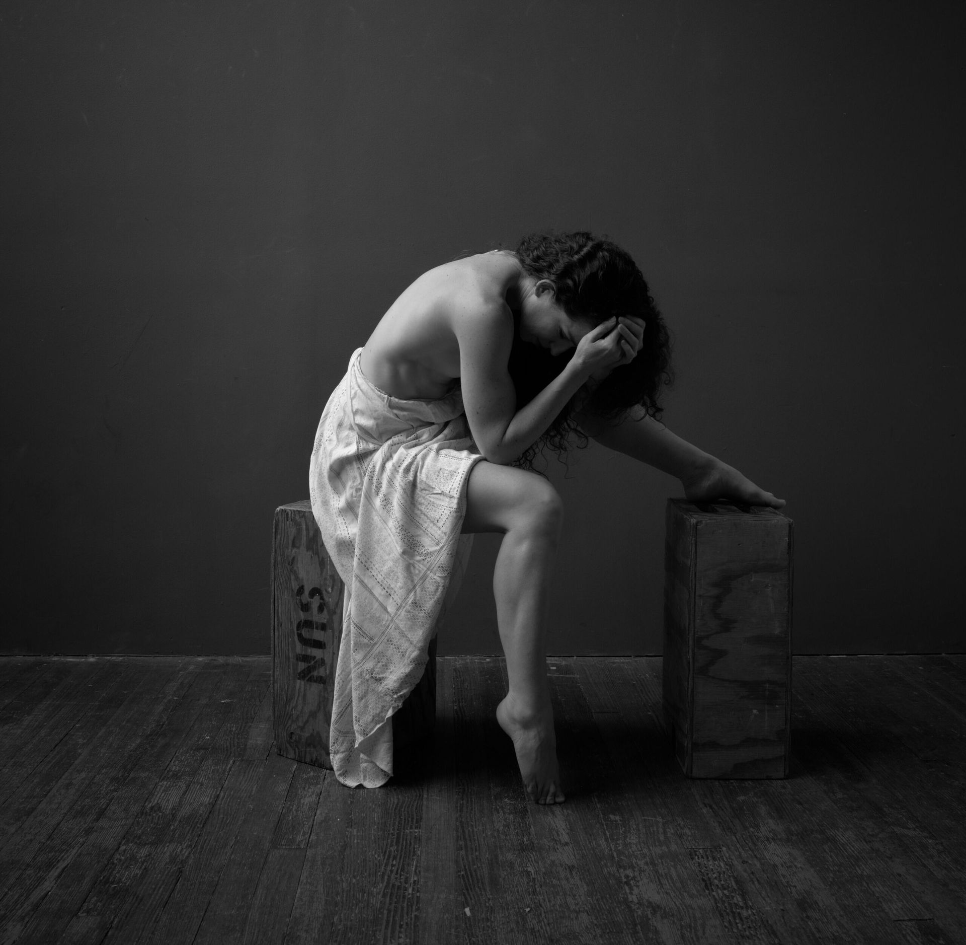 Une femme est assise sur une caisse en bois, la tête dans les mains, enveloppée dans un tissu noir et blanc.