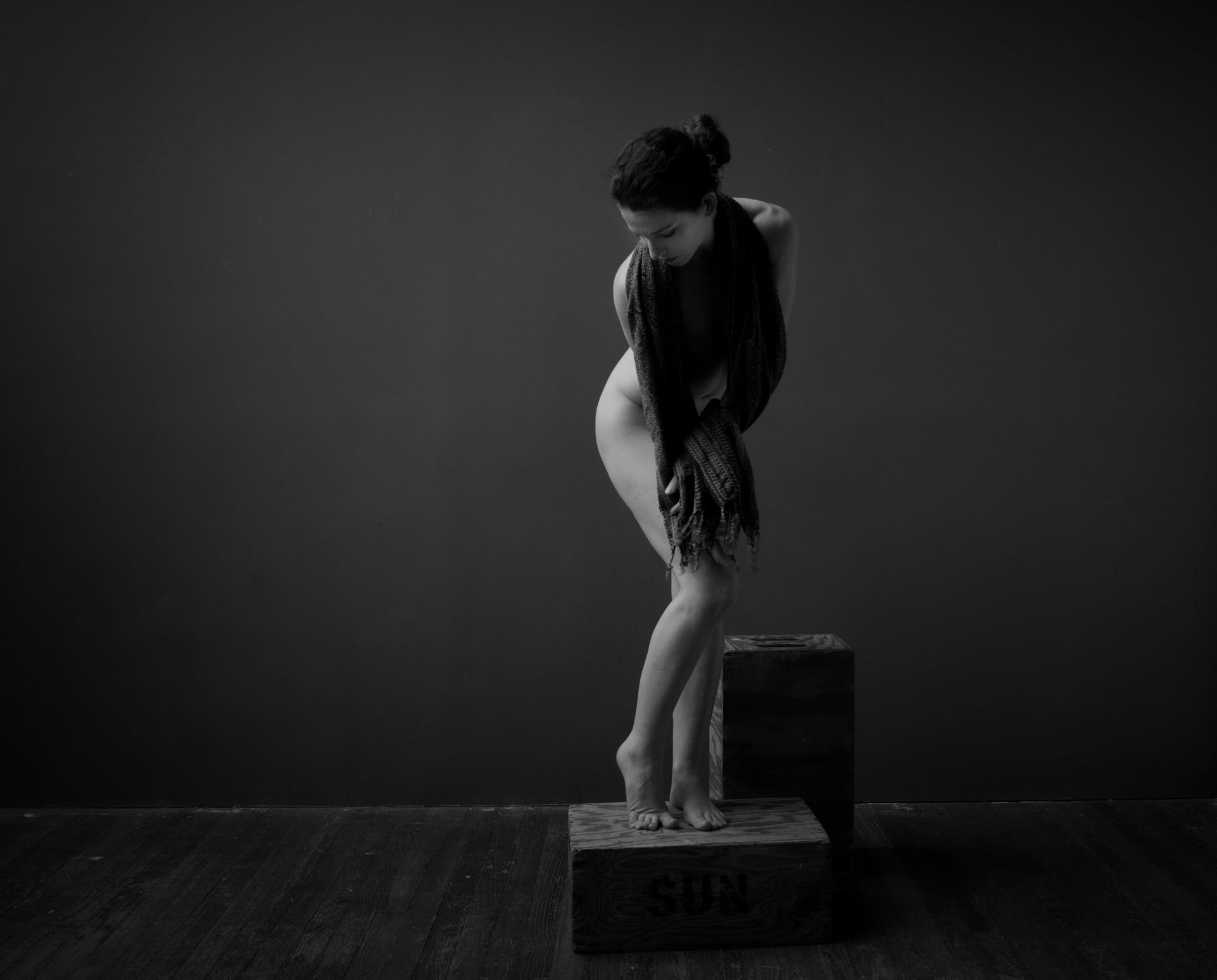 Figure nue debout sur une boîte, drapée de tissu, dans un studio à fond sombre.