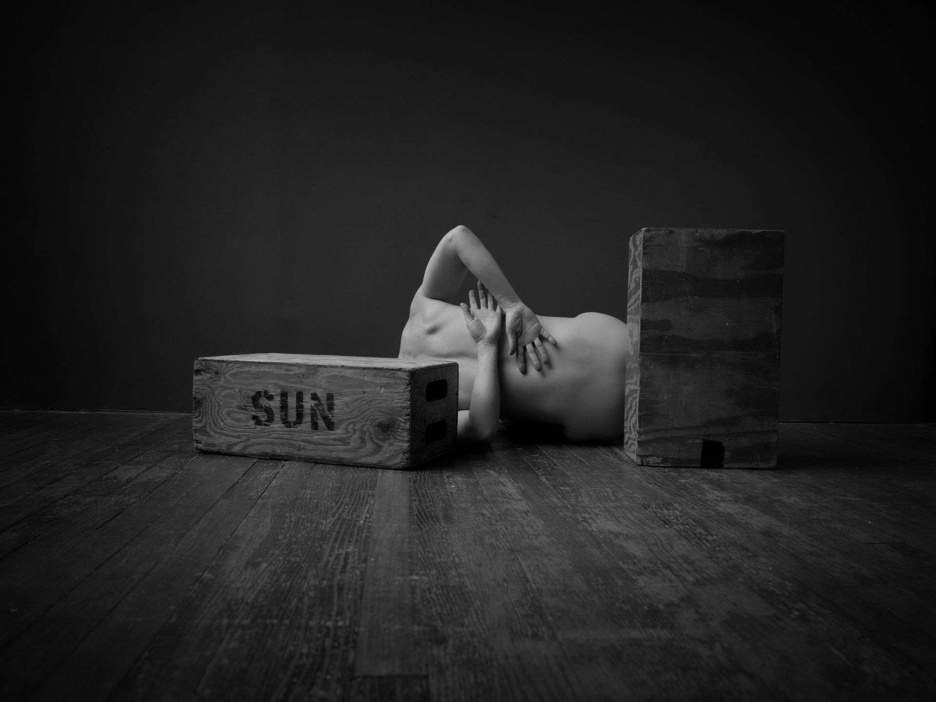 Personne nue allongée au sol, dos à l'objectif, briques avec l'inscription « SOLEIL » devant. Photo en noir et blanc.