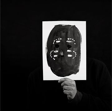Personne tenant une feuille de papier blanc avec un masque peint en noir, sur fond sombre.