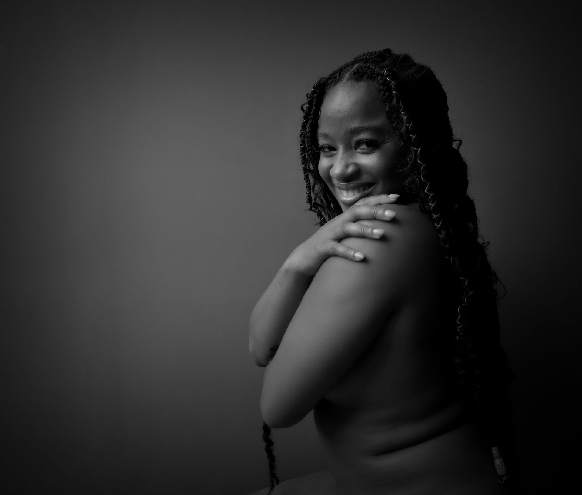 Personne souriante aux longues tresses, nue, se tenant l'épaule, sur fond gris.