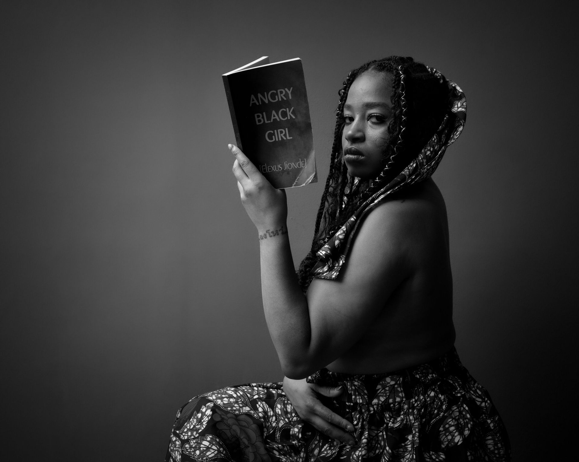 Femme vêtue d'un tissu à motifs, tenant un livre intitulé « La fille noire en colère ». Noir et blanc, assise, expression pensive.