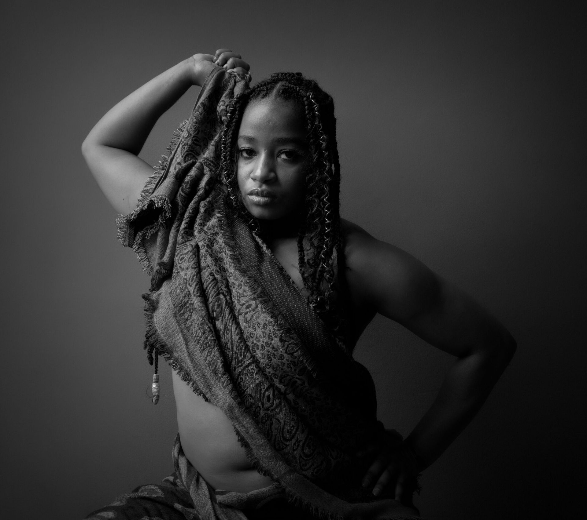 Femme drapée dans un tissu, le tenant au-dessus de sa tête. Cheveux tressés, regard fixé sur l'objectif. Noir et blanc.