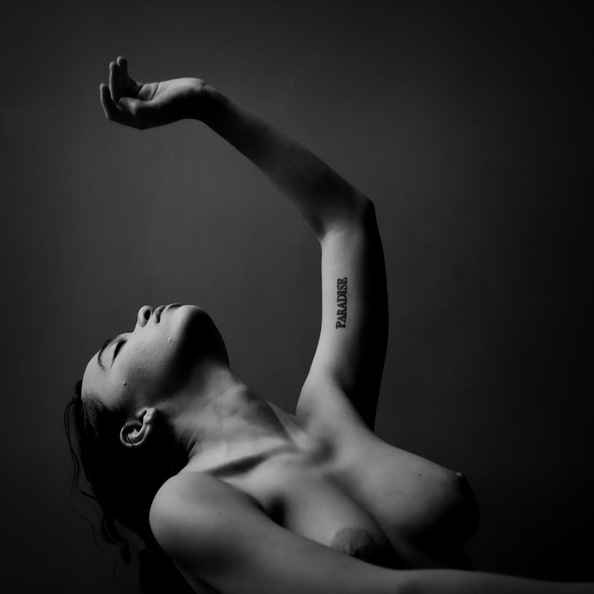 Femme, bras levé, tête renversée. Noir et blanc, tatouage visible sur le haut du bras.