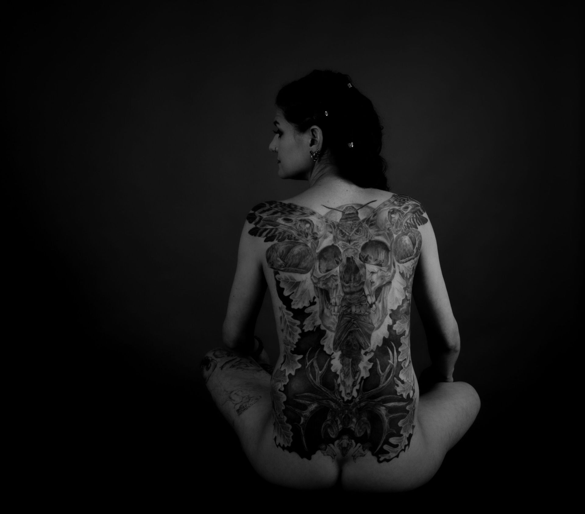 Une femme au dos entièrement tatoué est assise en tailleur, dos au spectateur, dans un studio faiblement éclairé.