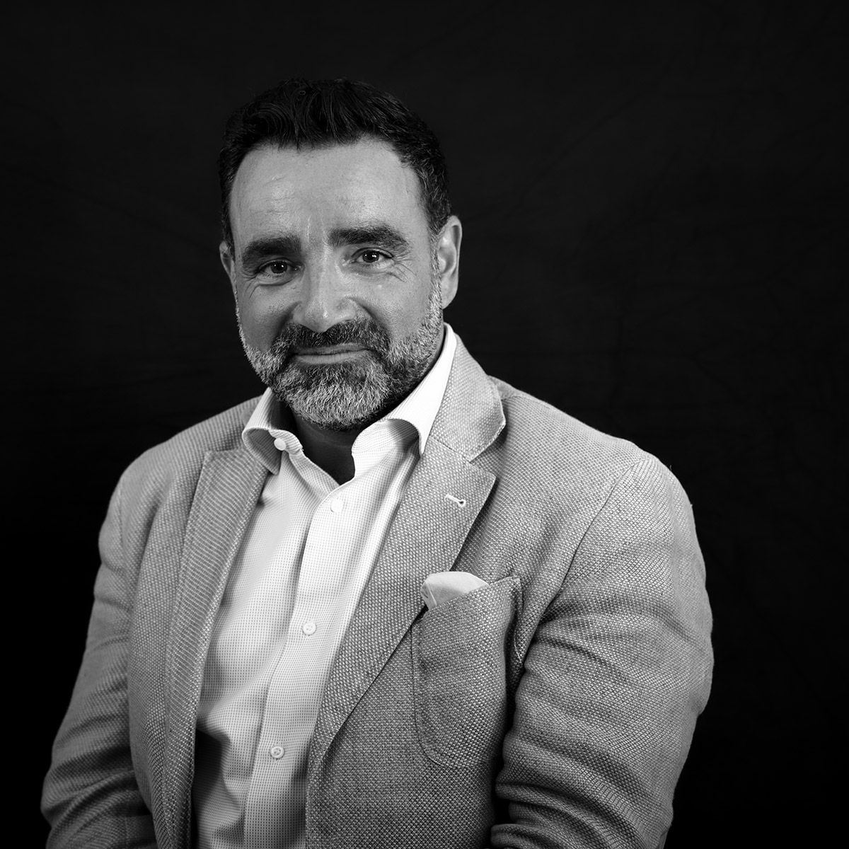 Homme en blazer et chemise à col ouvert, souriant, sur fond sombre.