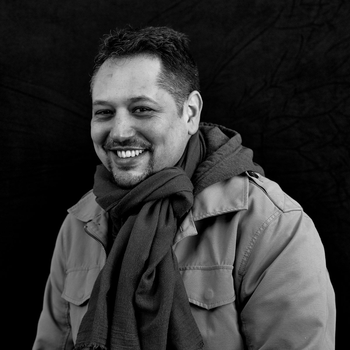 Un homme souriant, portant une écharpe et une veste, se détache sur un fond sombre.