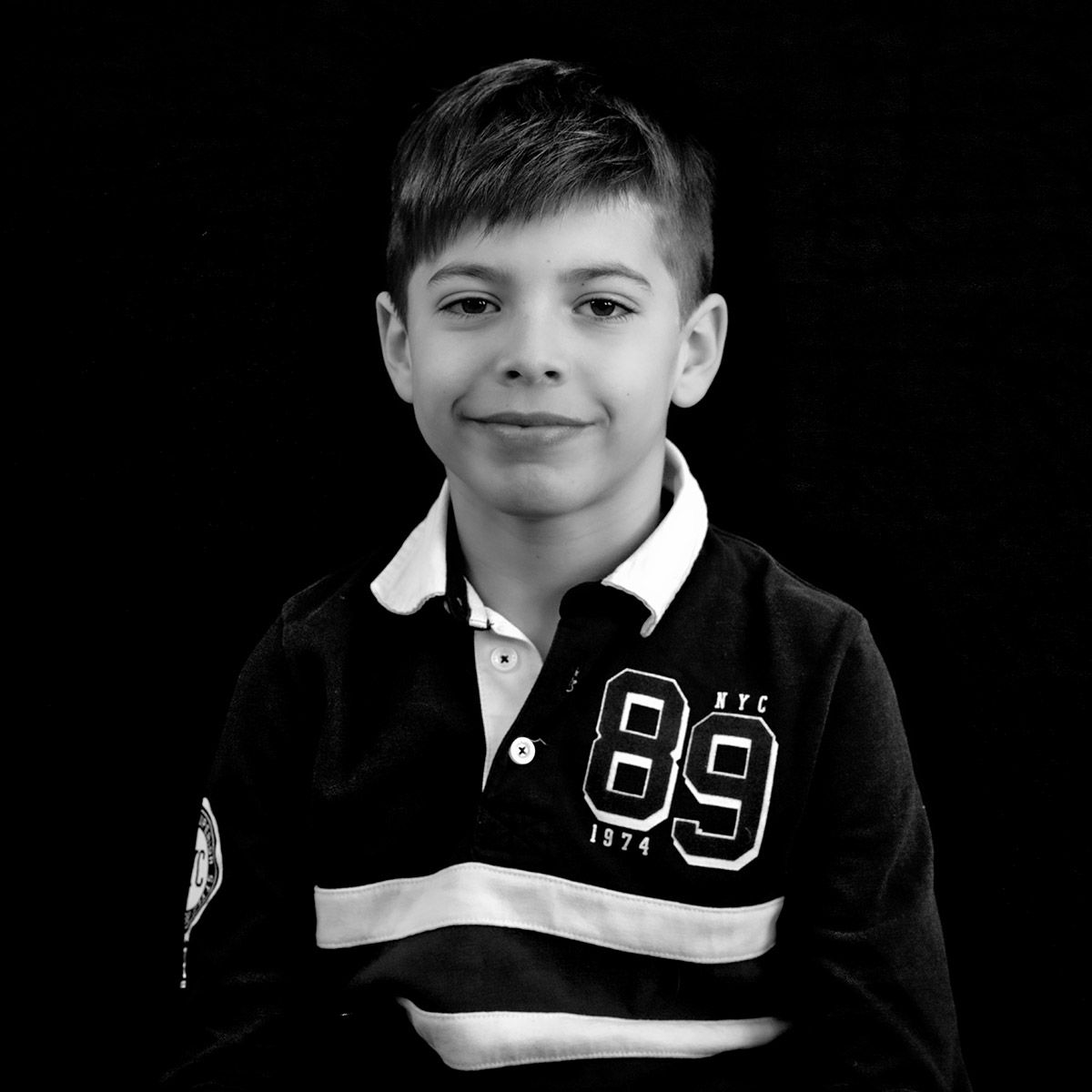 Garçon portant un pull bleu foncé avec un écusson « 89 NYC », souriant à l'objectif.