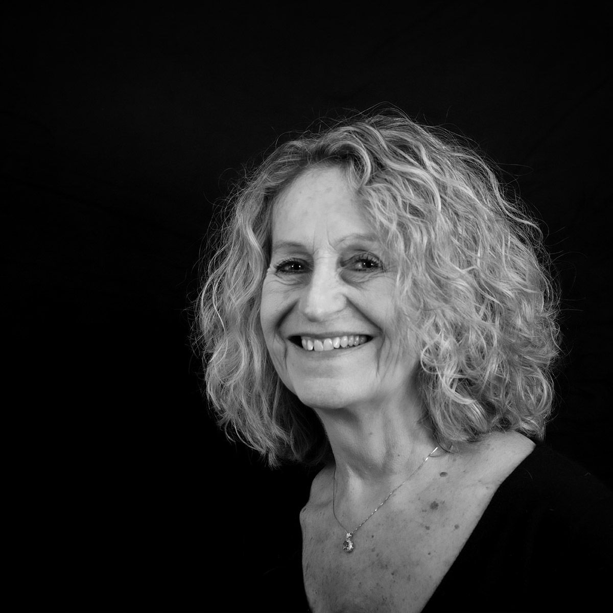 Une femme aux cheveux bouclés sourit, face à la caméra, sur fond noir.