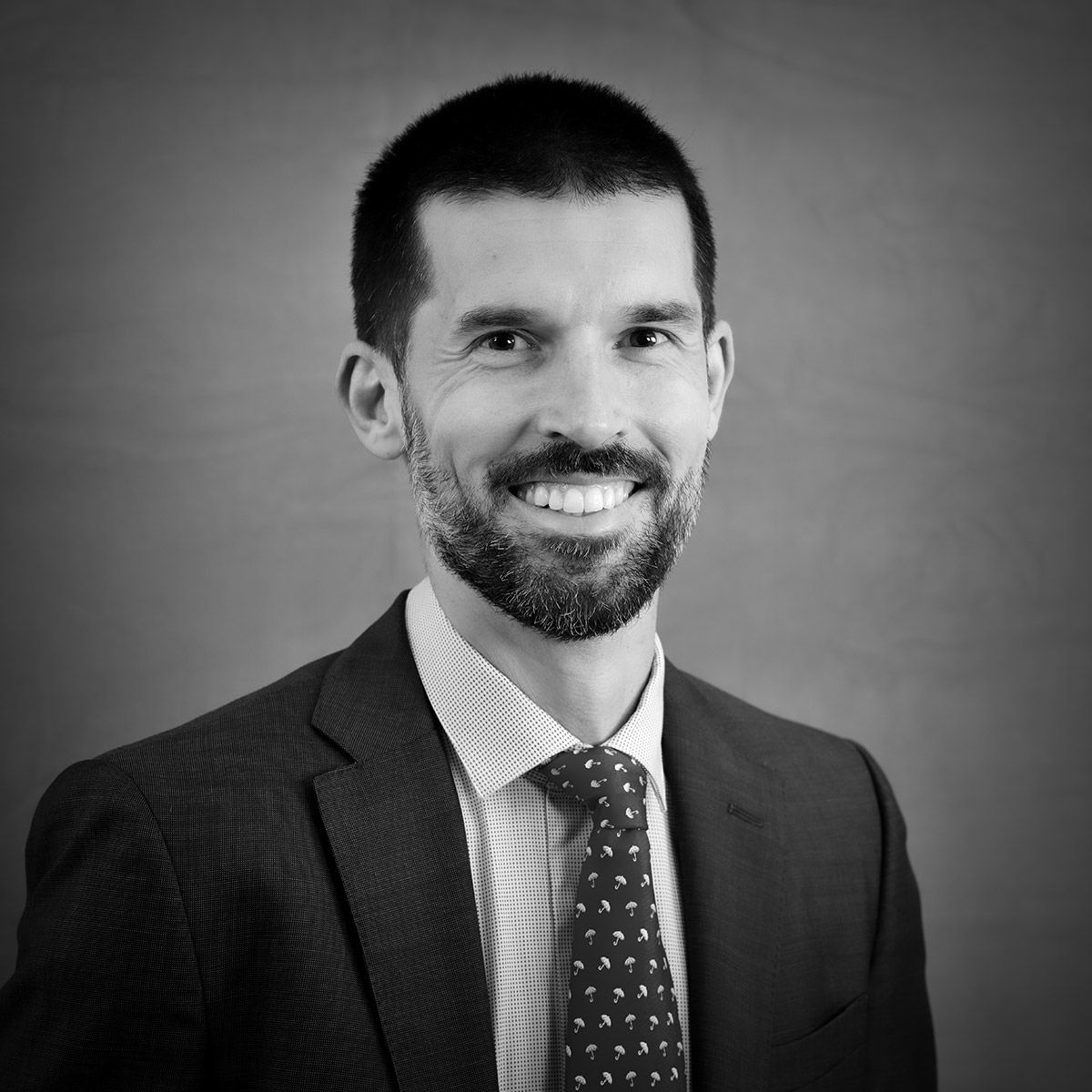 Un homme en costume sourit en regardant l'objectif. Cravate sombre, chemise à motifs et barbe soigneusement taillée.