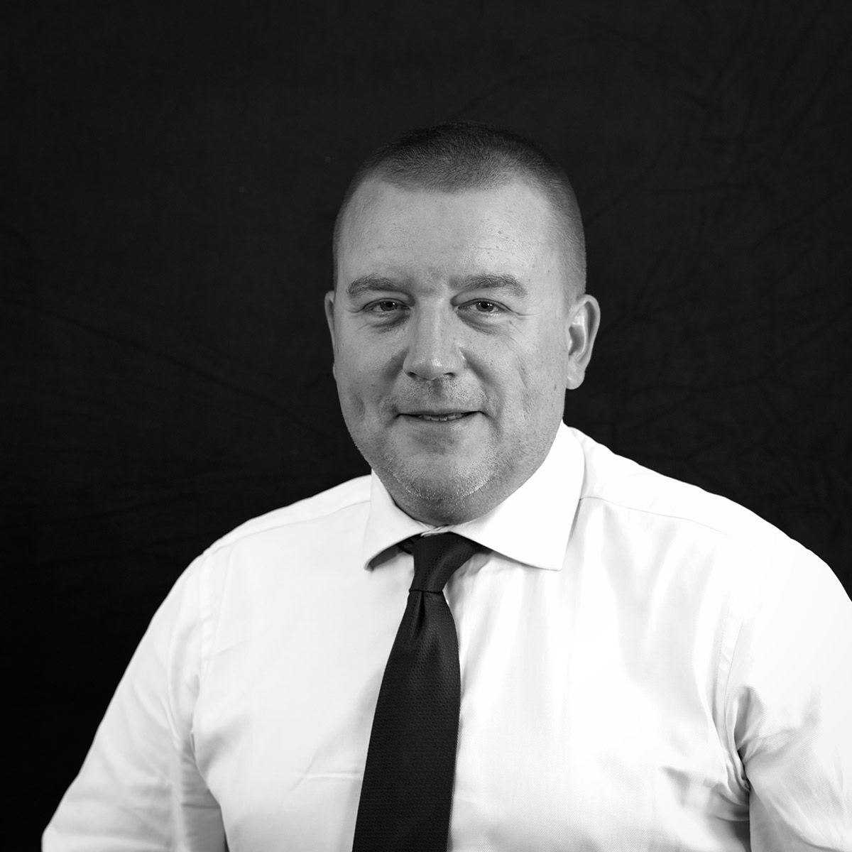 Homme en chemise blanche et cravate sur fond sombre, esquissant un sourire.