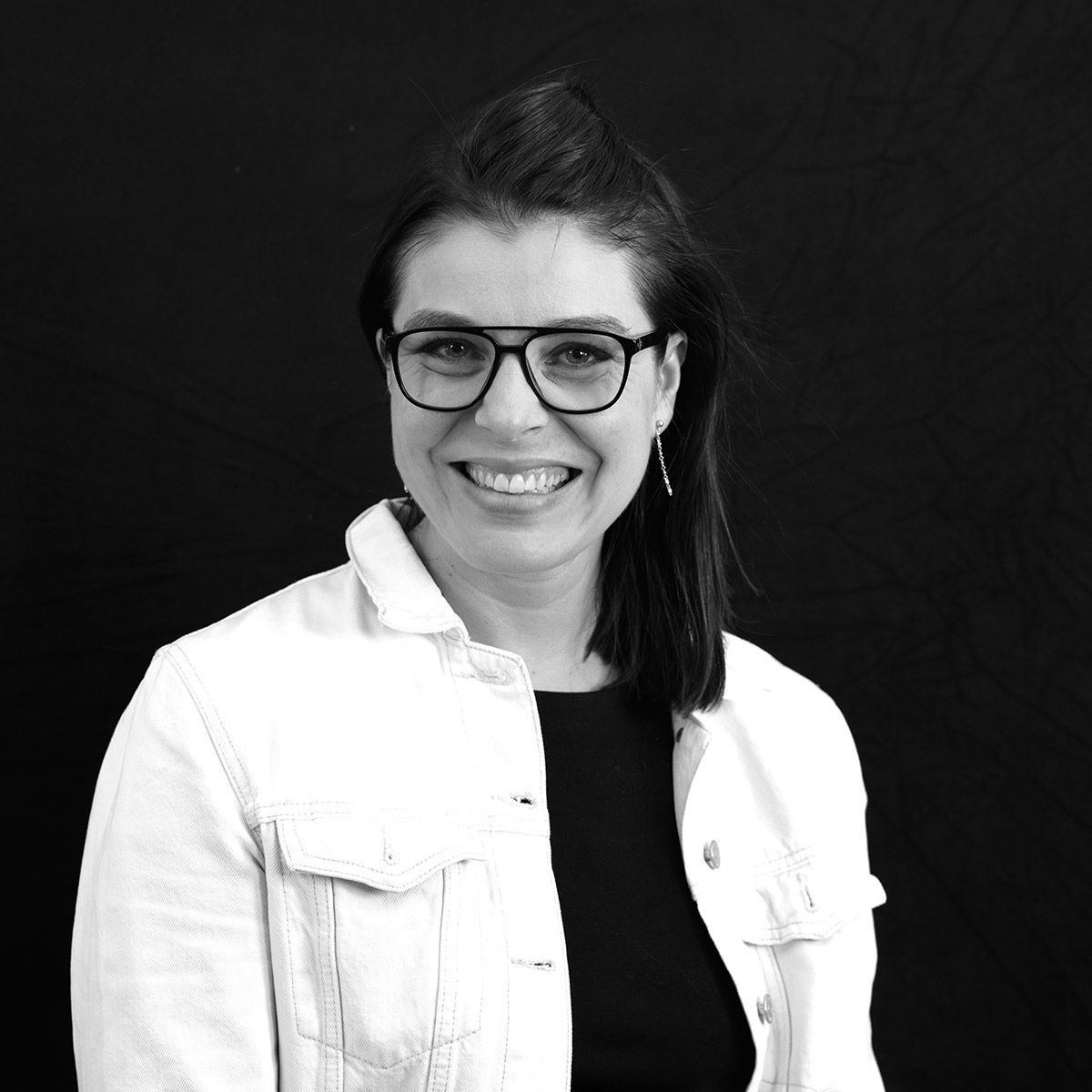 Femme à lunettes, souriante, portant une veste blanche sur un haut noir, sur fond noir.