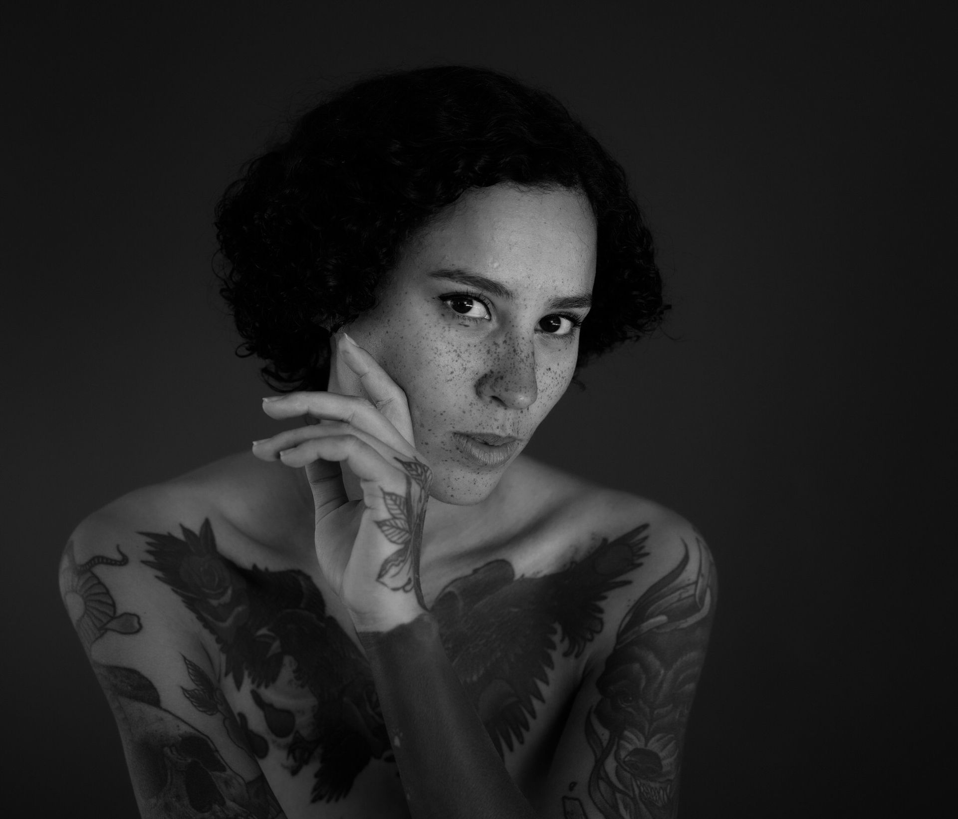 Femme aux cheveux bouclés et tatouée, la main au visage, regardant le spectateur. Noir et blanc.