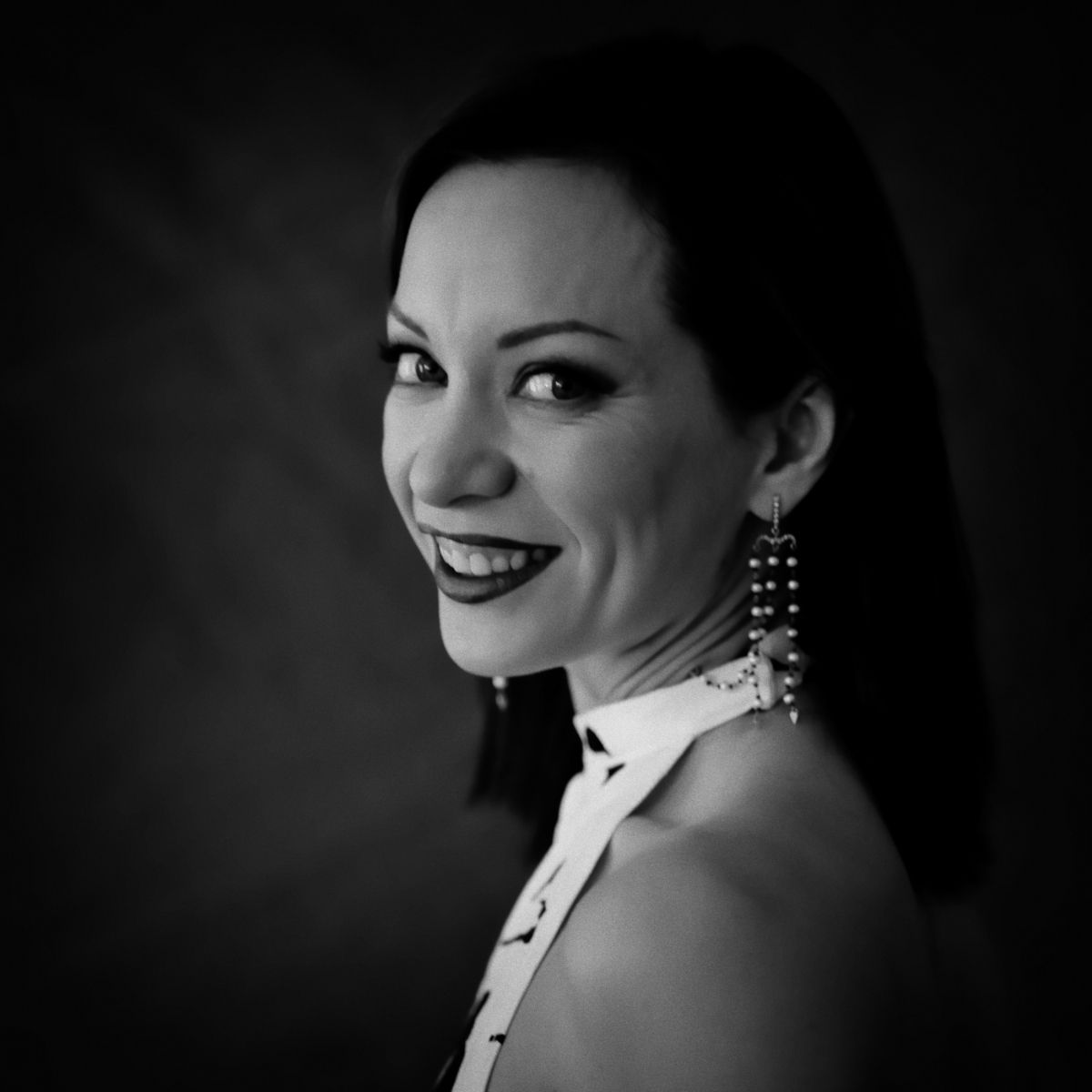 Femme aux cheveux foncés, souriante, portant des boucles d'oreilles, regardant par-dessus son épaule, sur fond sombre.