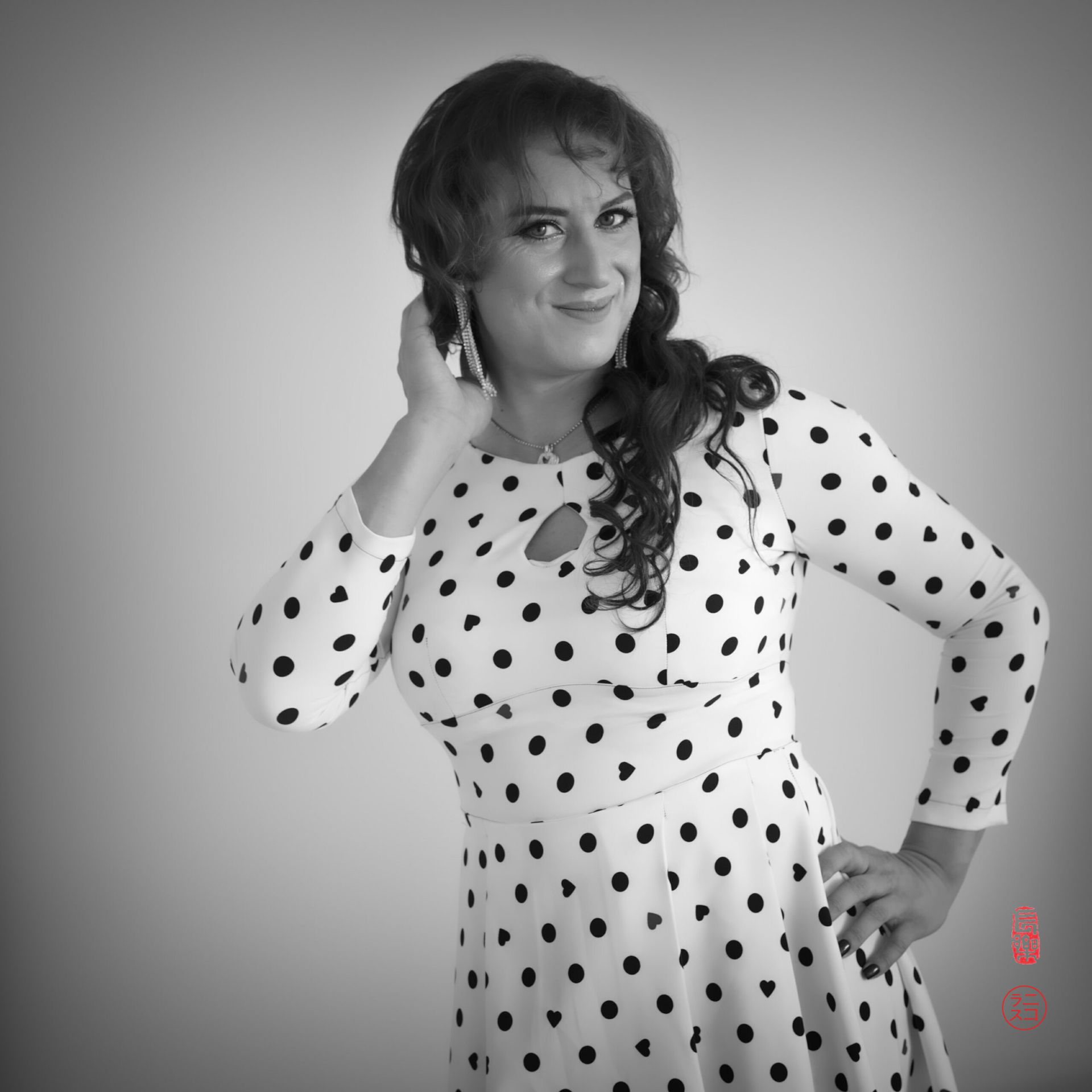 Femme en robe à pois, souriante, main dans les cheveux. Décor de studio.