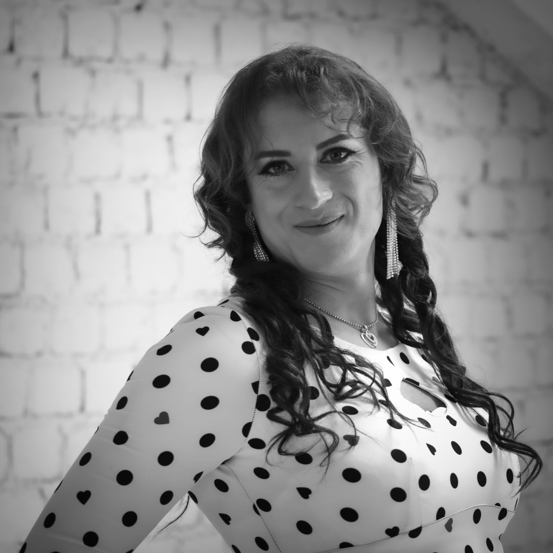 Femme aux longs cheveux noirs et bouclés, vêtue d'une robe à pois, souriant à l'objectif, debout devant un mur de briques blanches.