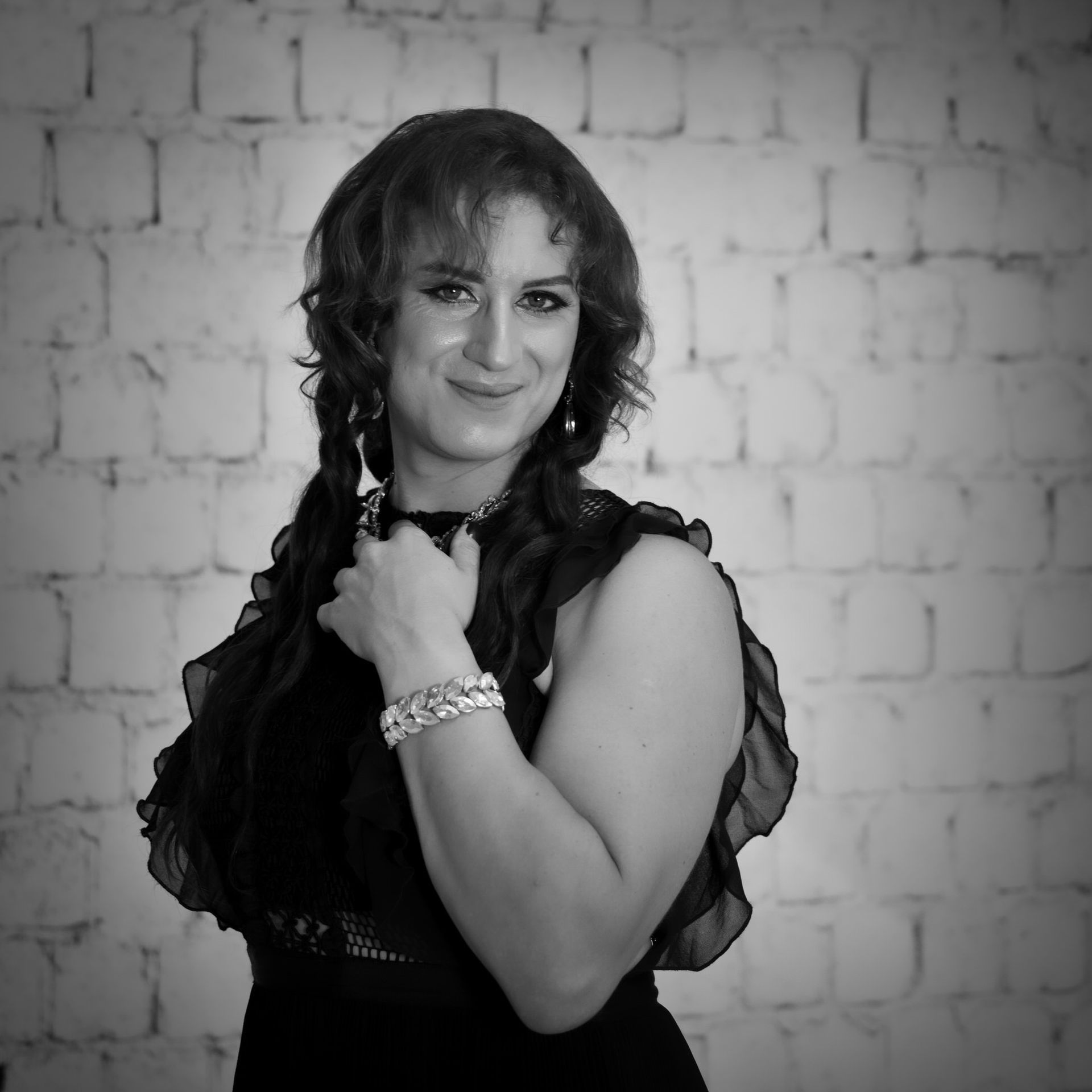 Femme aux cheveux bouclés tressés en nattes, souriante et tenant son poignet, devant un mur de briques blanches.