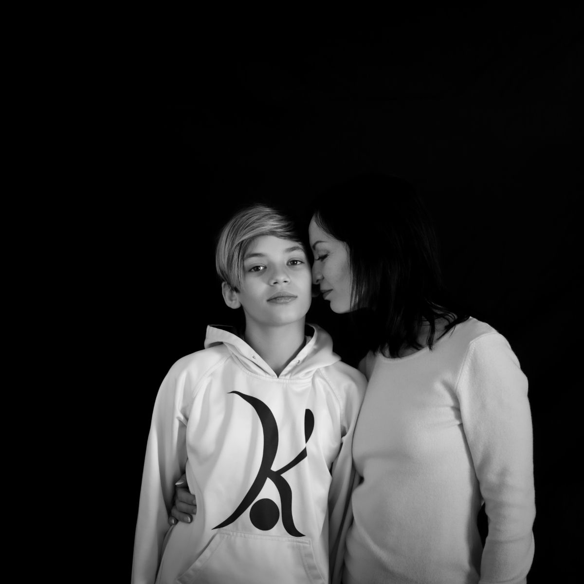 Une femme chuchote à une personne portant un sweat à capuche, toutes deux sur fond noir.