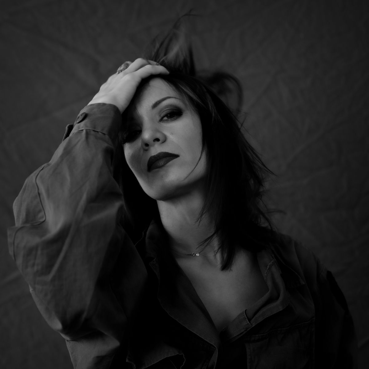 Femme en chemise sombre, main dans les cheveux, regardant vers le ciel avec une expression neutre. Photo en noir et blanc.