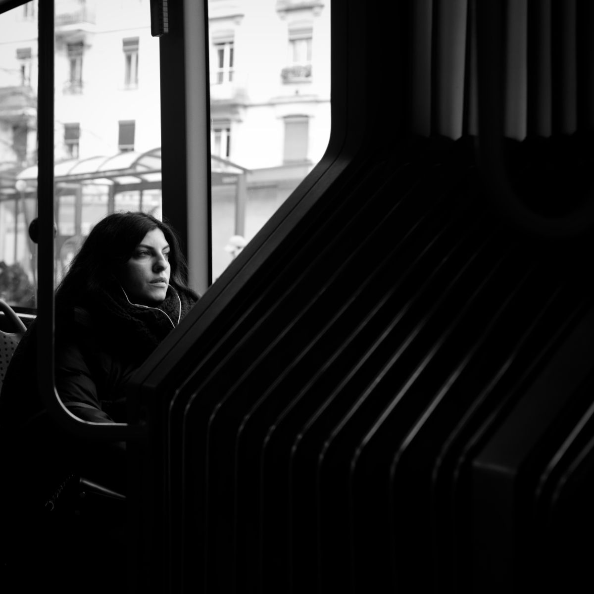 Une femme dans un bus regarde par la fenêtre, son visage est ombragé par les sièges. On aperçoit des immeubles à travers la vitre.