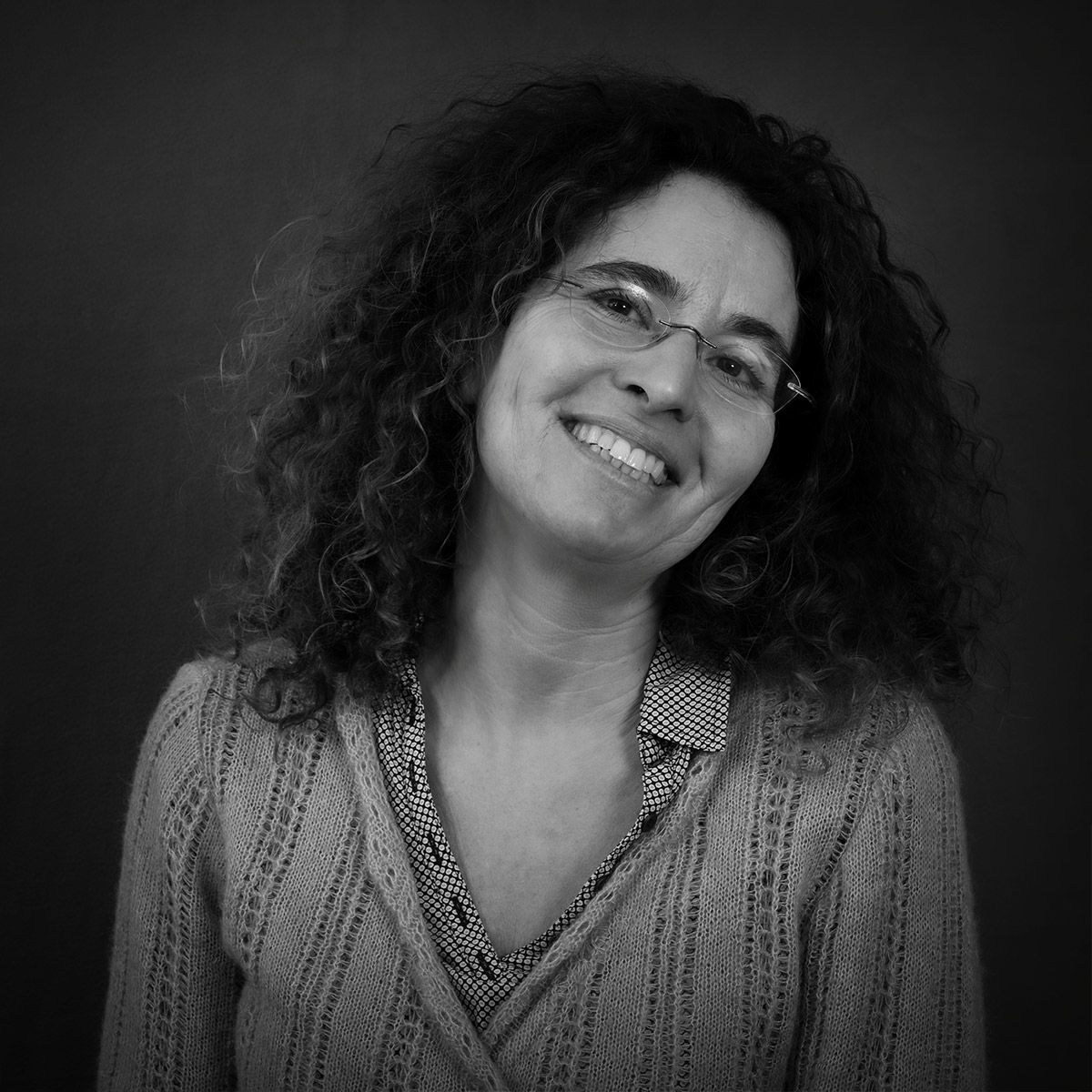 Femme aux cheveux bouclés, souriante, portant des lunettes et un pull à motifs, sur fond sombre.