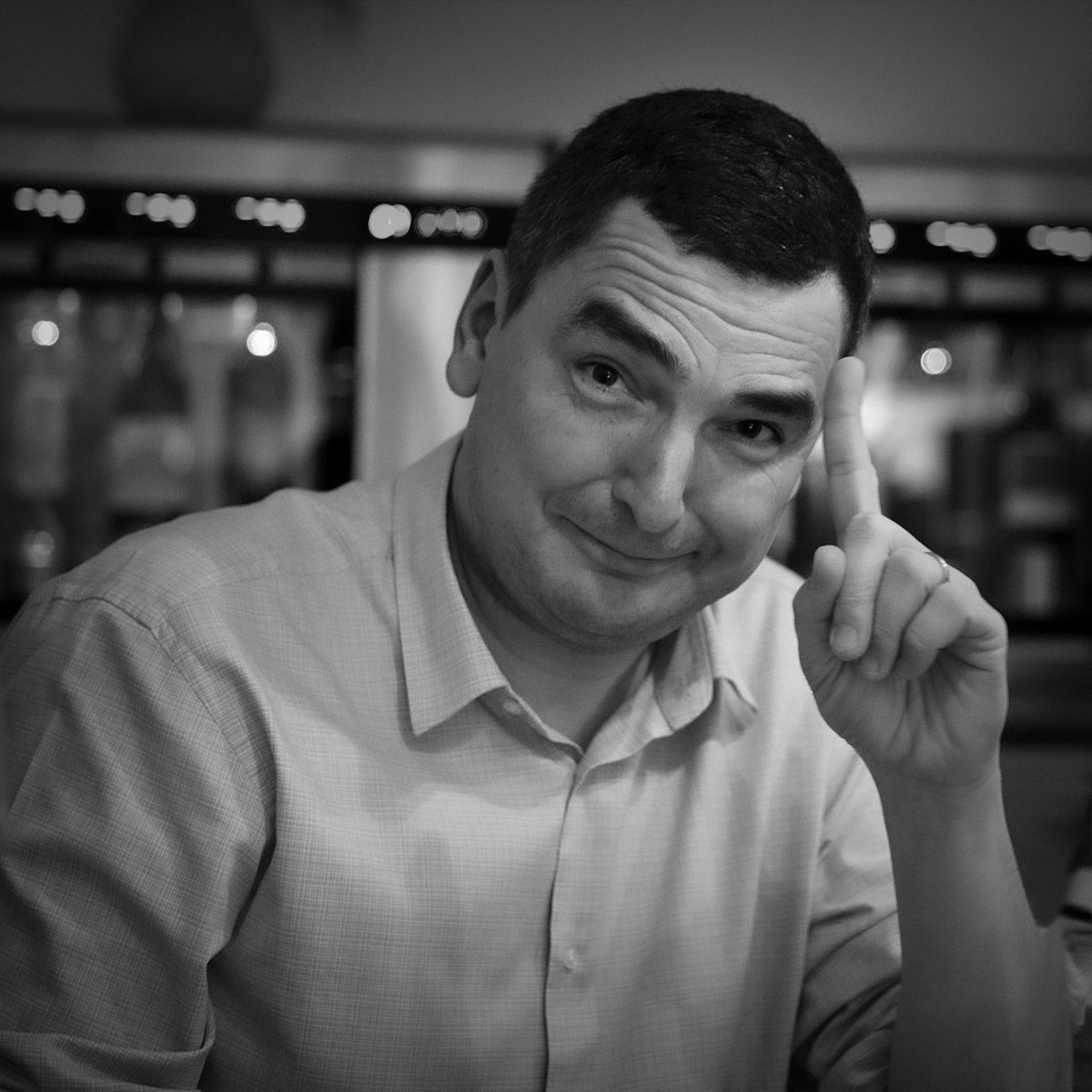 Un homme aux cheveux foncés pointe du doigt vers le haut en souriant. Il porte une chemise claire à boutons, à l'intérieur.