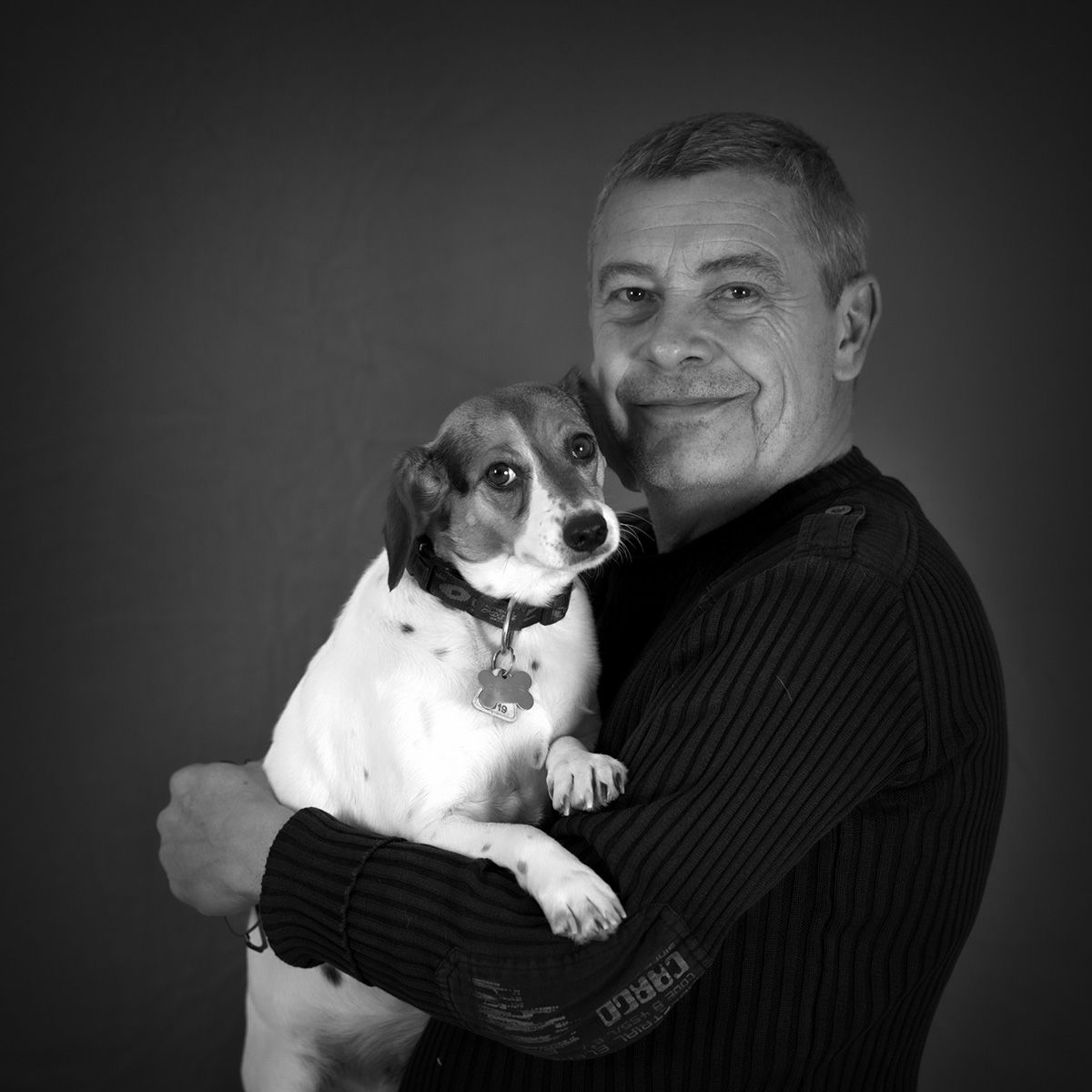 Un homme tenant un petit chien, tous deux souriant à l'objectif. Noir et blanc, en studio.