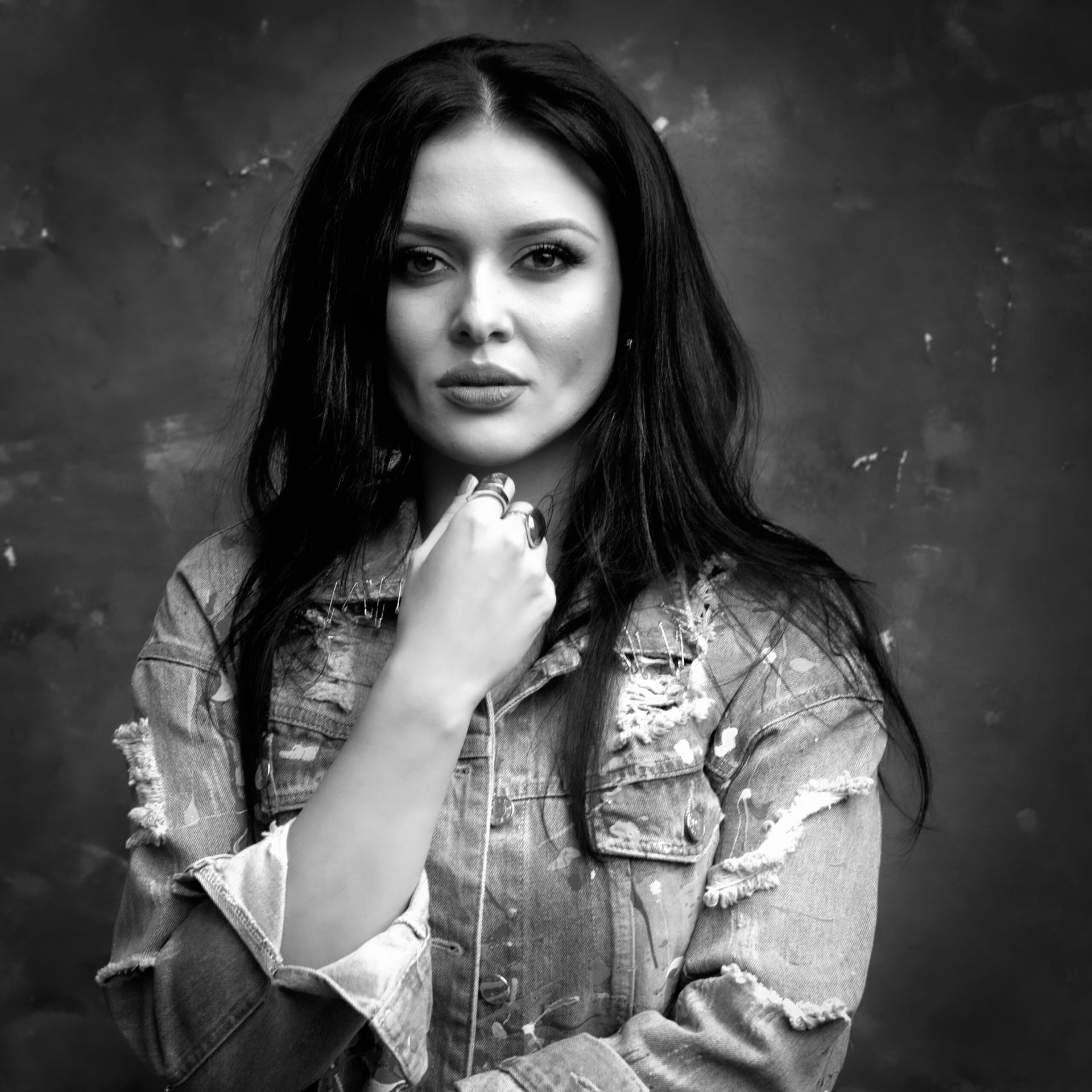 Femme en veste en jean délavée, la main près du visage, regardant le spectateur. Portrait en noir et blanc sur fond moucheté.