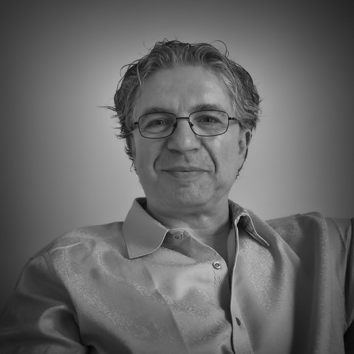 Un homme portant des lunettes et une chemise boutonnée sourit pour un portrait, éclairage doux, niveaux de gris.