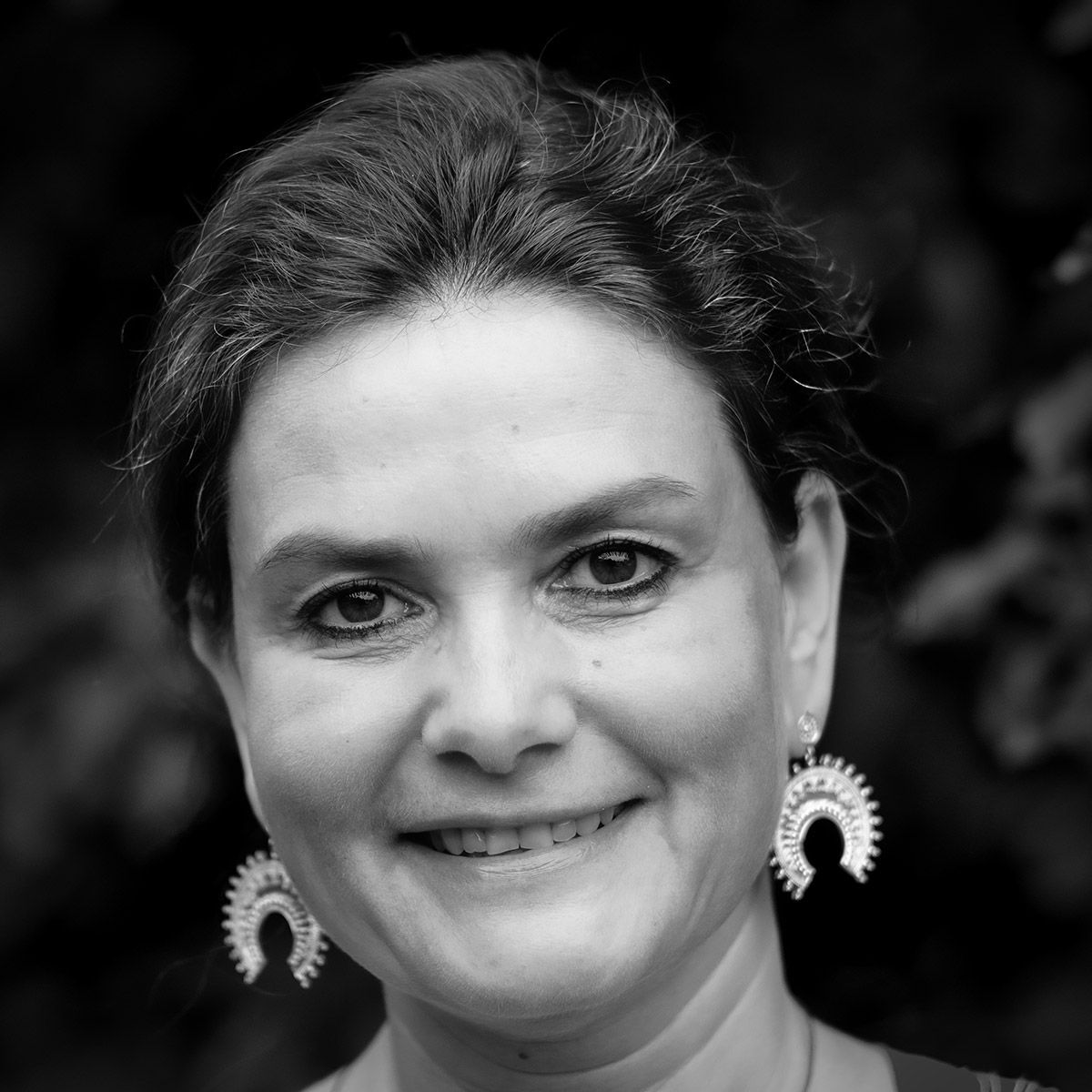 Une femme aux cheveux courts sourit, portant des boucles d'oreilles décoratives, en extérieur.