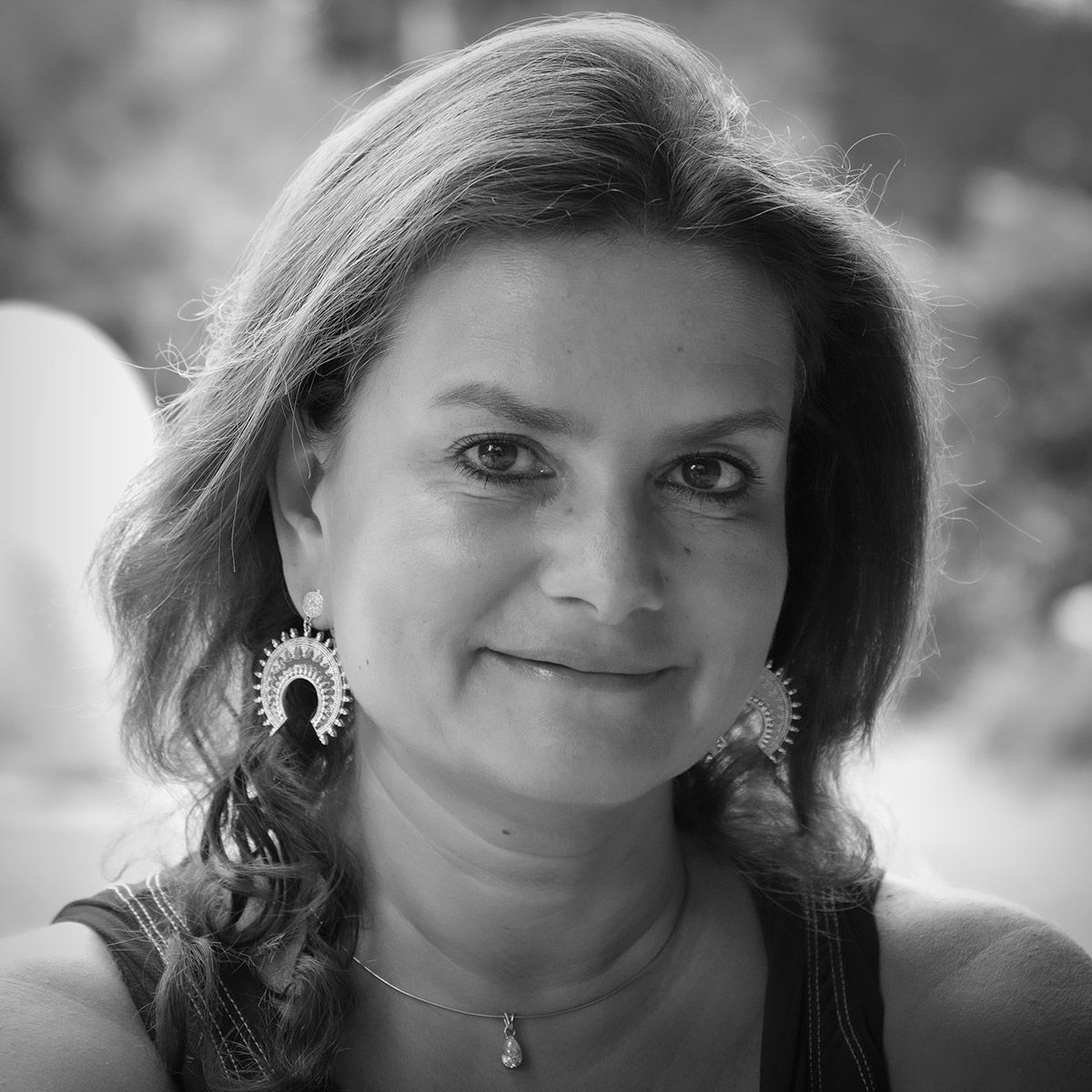 Femme aux cheveux bruns mi-longs, souriante, portant de grandes boucles d'oreilles pendantes et un collier pendentif.