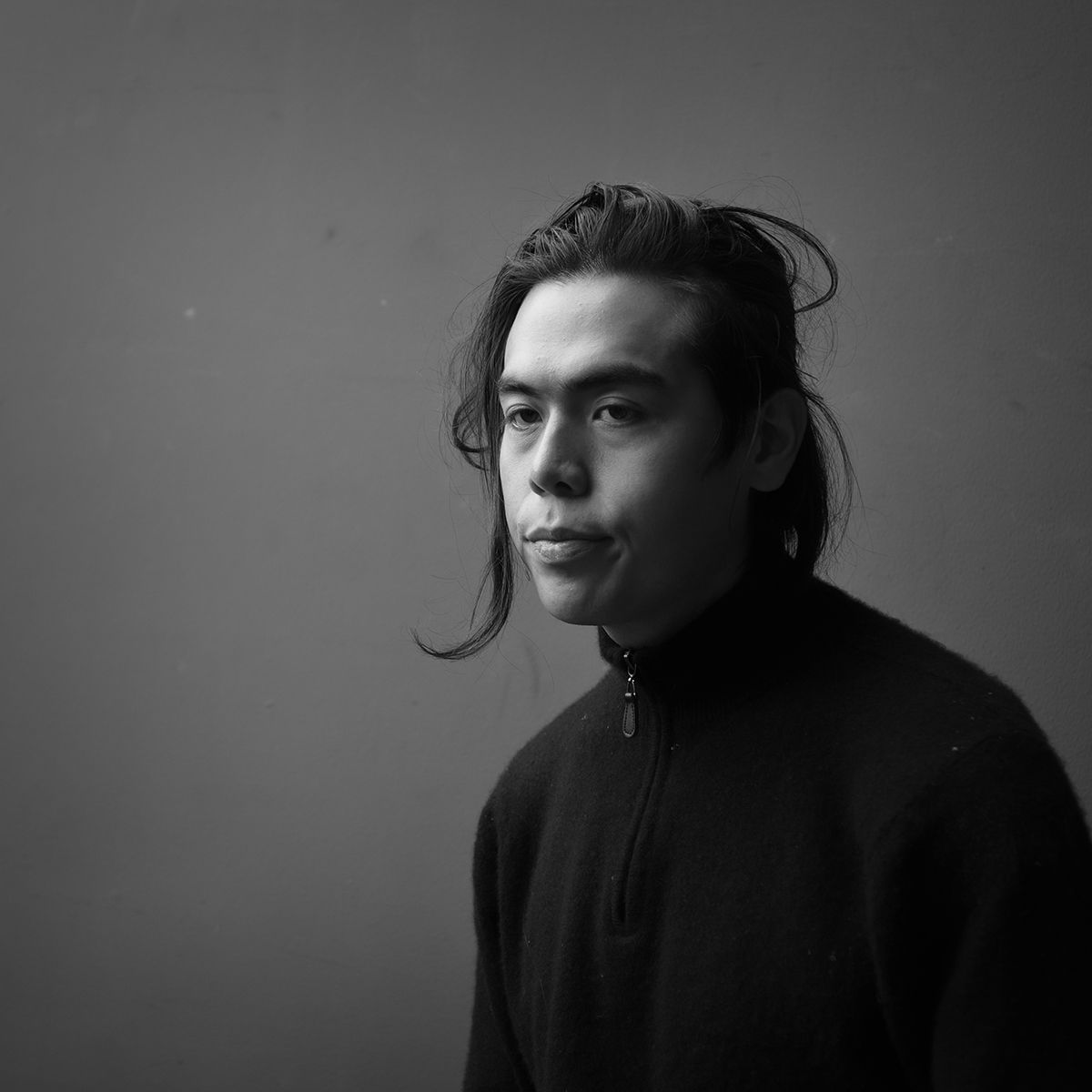 Homme aux cheveux longs, portant un pull foncé, regardant sur le côté sur une photo de studio en noir et blanc.