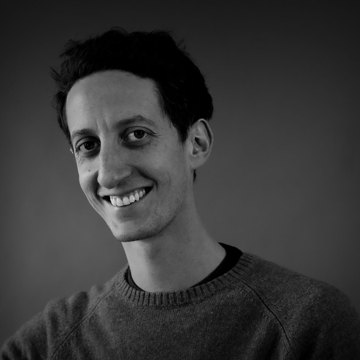 Un homme souriant aux cheveux foncés, vêtu d'un pull gris, regarde l'objectif.