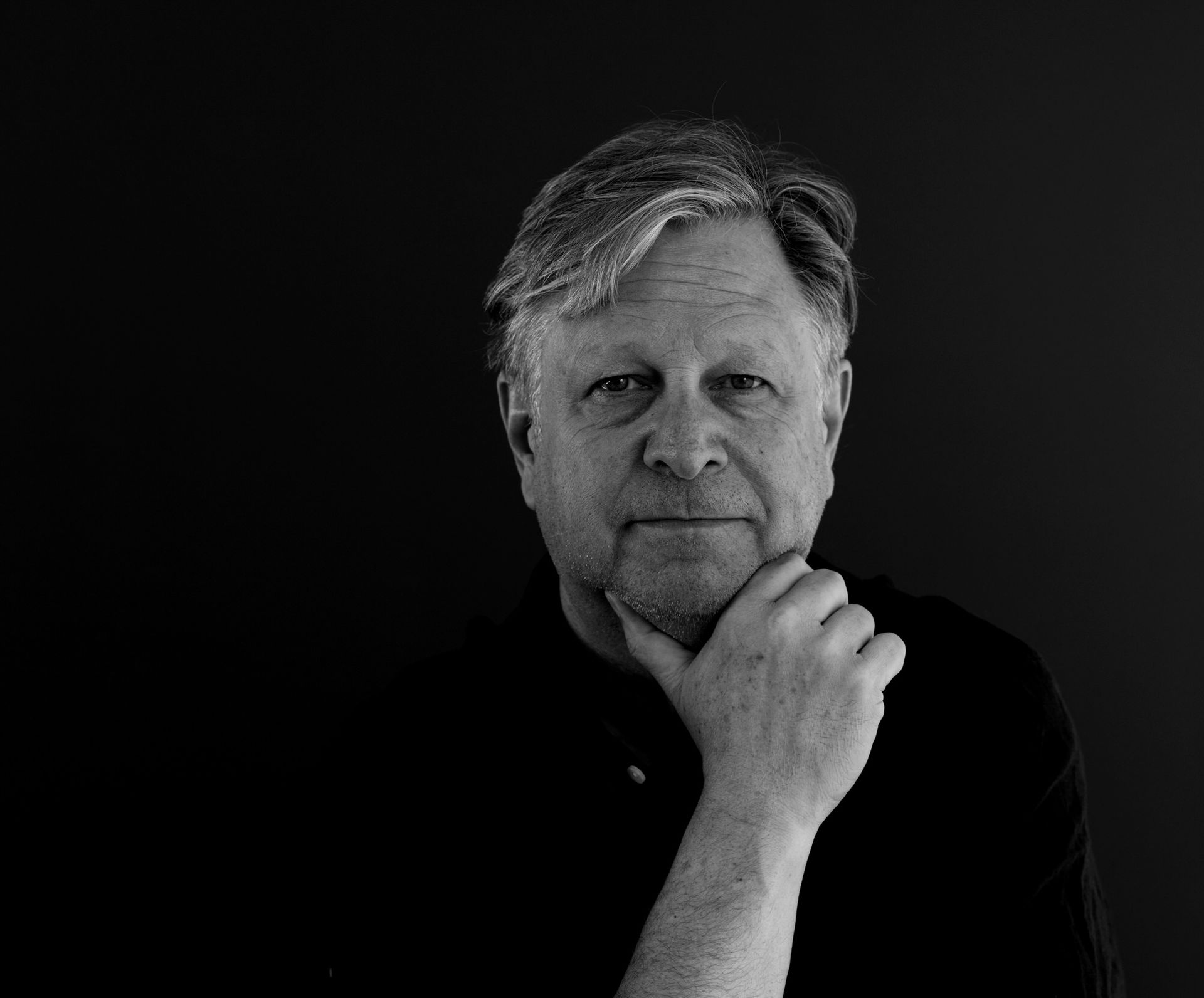 Homme aux cheveux gris courts, la main sur le menton, regardant pensivement l'objectif. Noir et blanc.