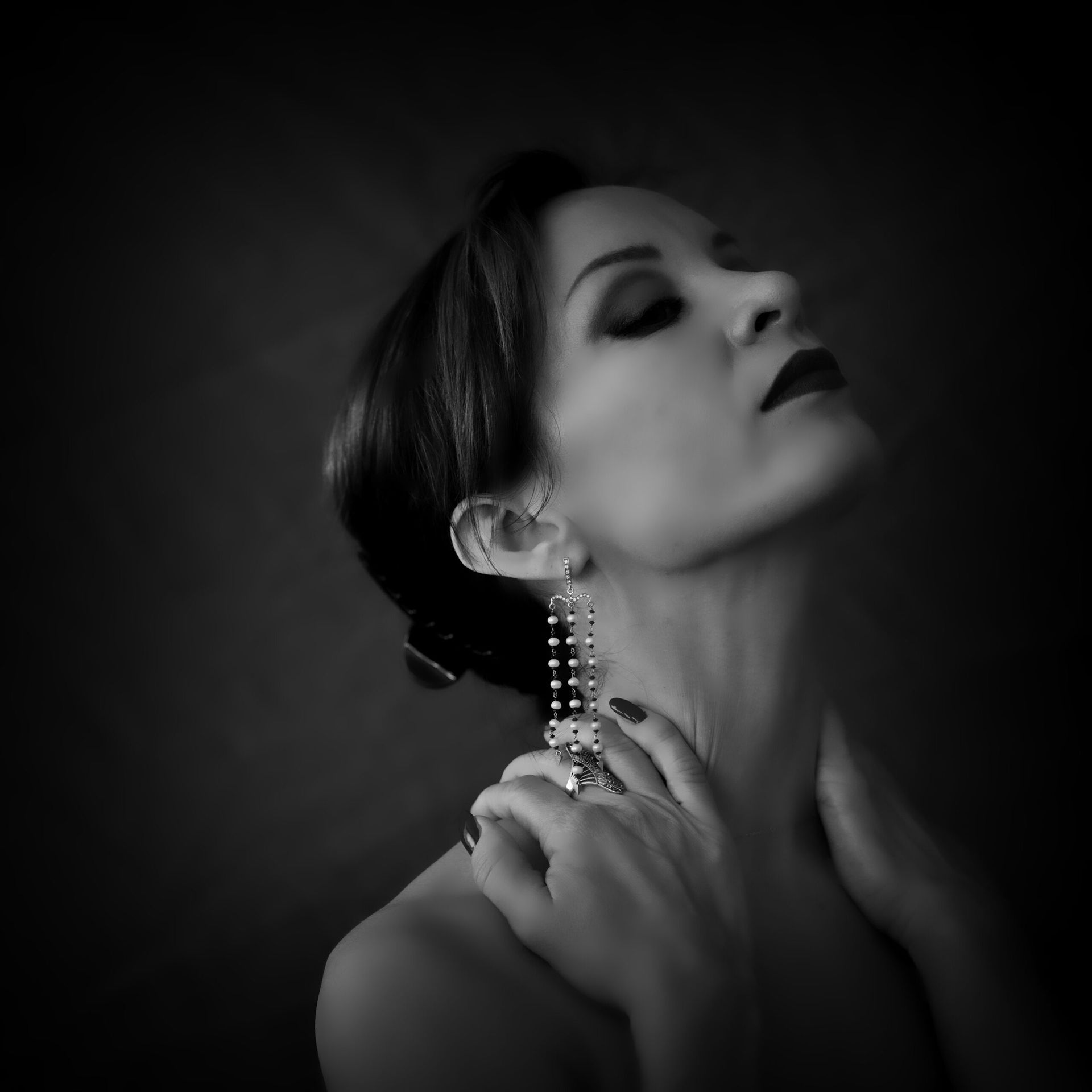 Femme portant des boucles d'oreilles, la tête renversée en arrière, les mains près du cou, maquillage foncé, noir et blanc.