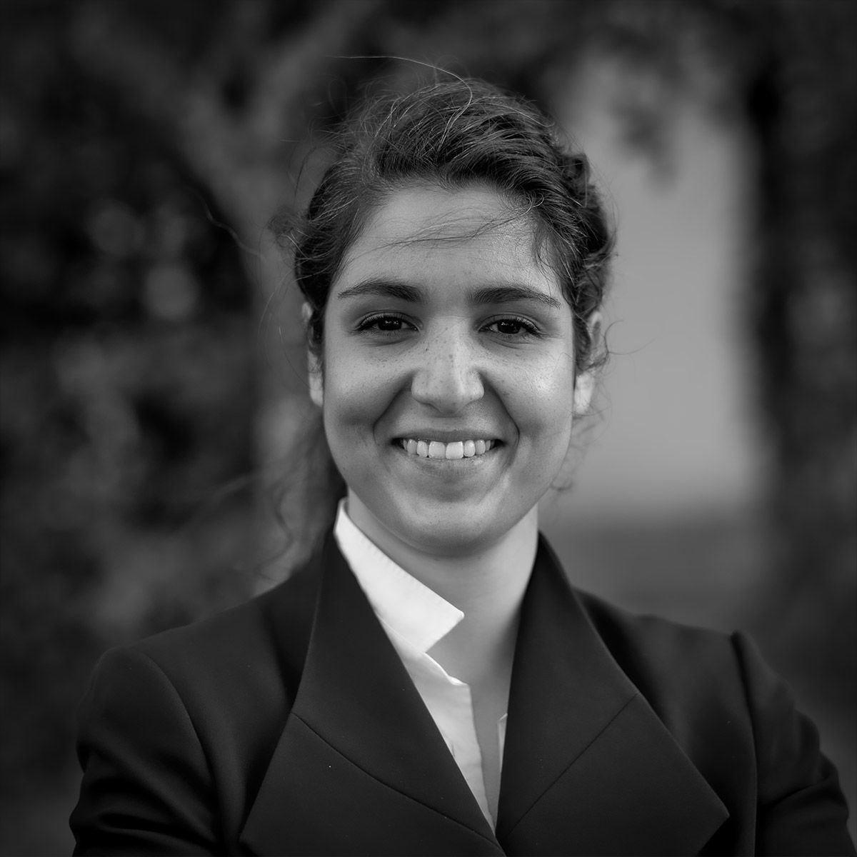 Femme en veste de tailleur souriante, en extérieur, avec des arbres en arrière-plan flou.