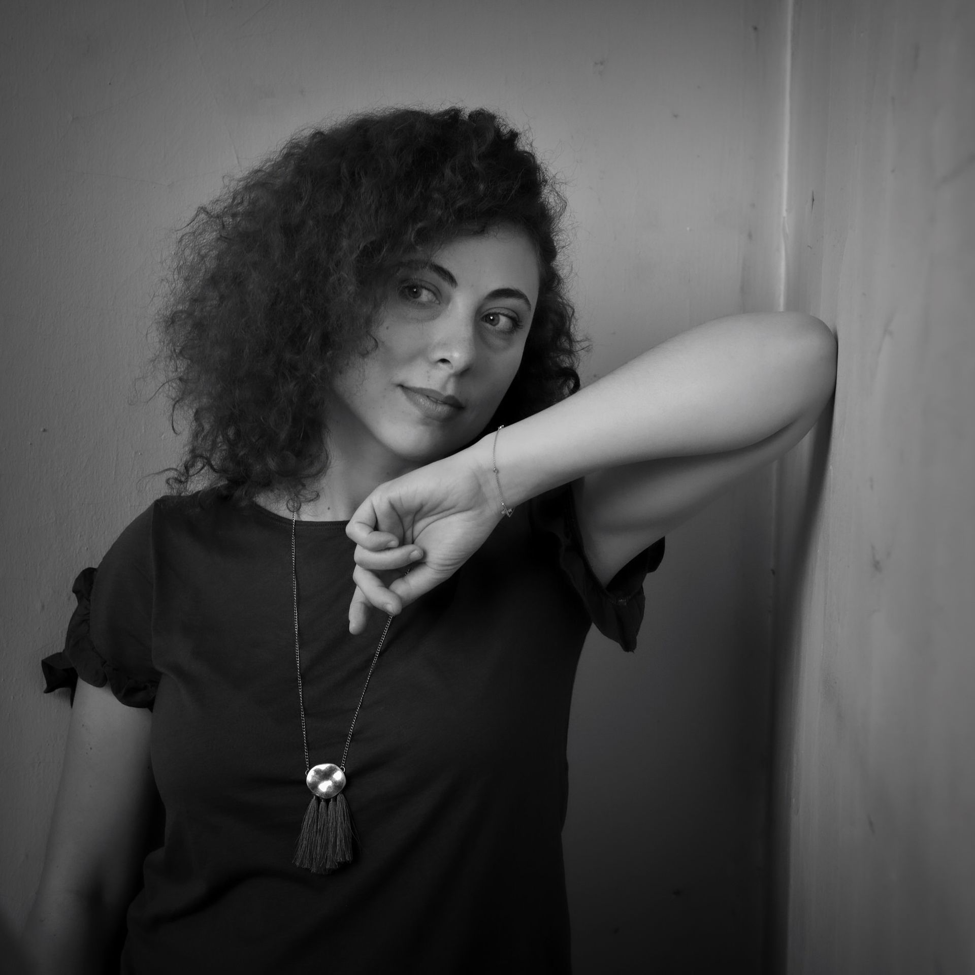 Une femme aux cheveux bouclés, le bras levé, est appuyée contre un mur. Elle porte une chemise sombre et un collier, et regarde le spectateur.