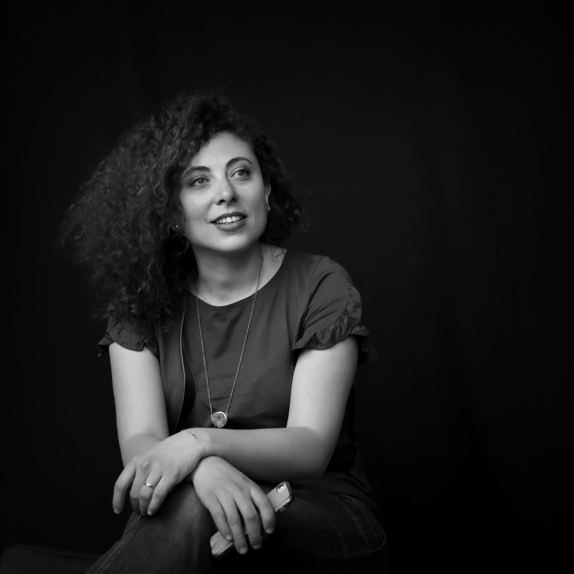 Femme aux cheveux bouclés, regardant vers le ciel et souriante, portant un collier et un haut foncé. Fond noir.