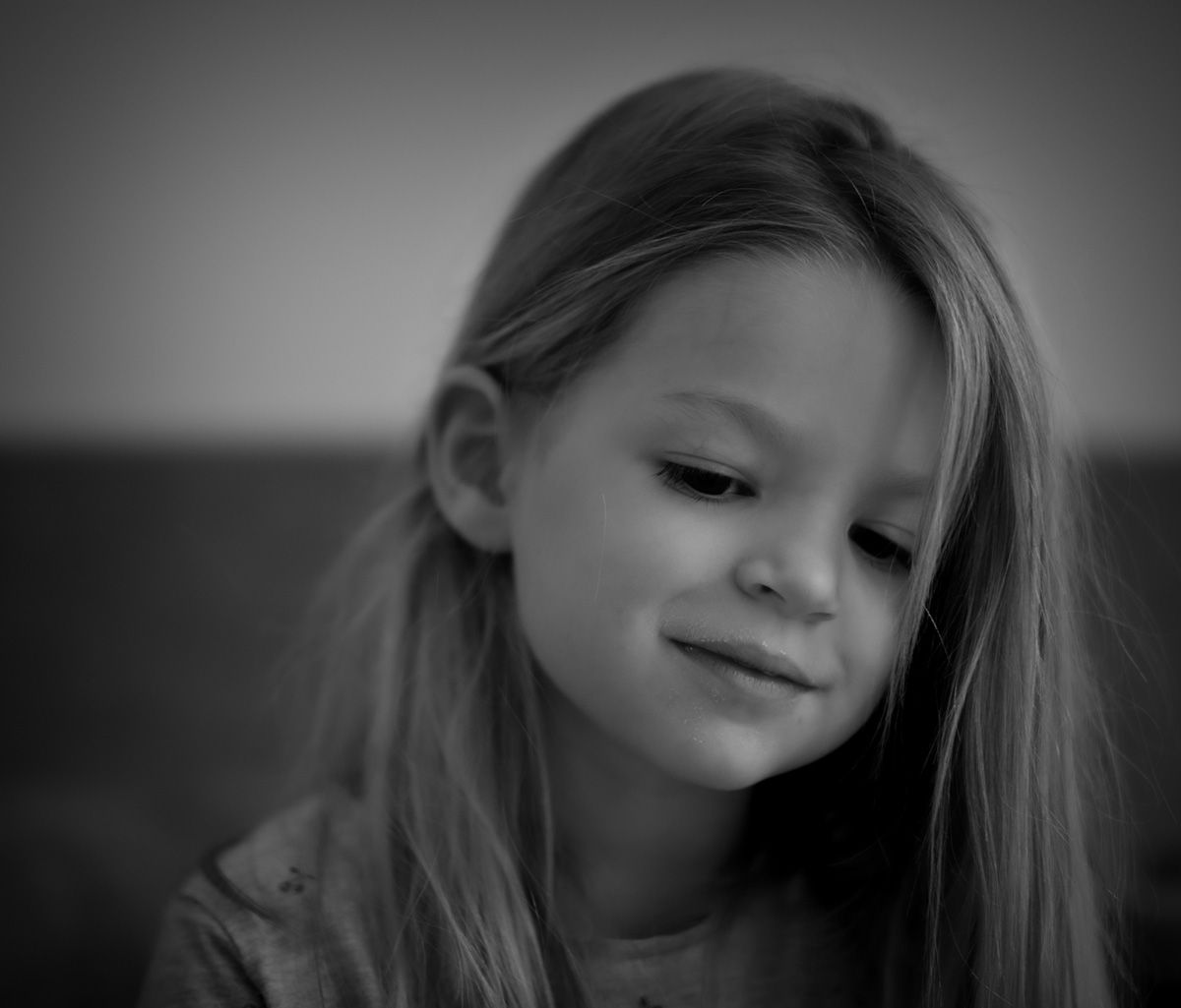 Enfant aux longs cheveux, le regard baissé, esquissant un sourire. Photo en noir et blanc.