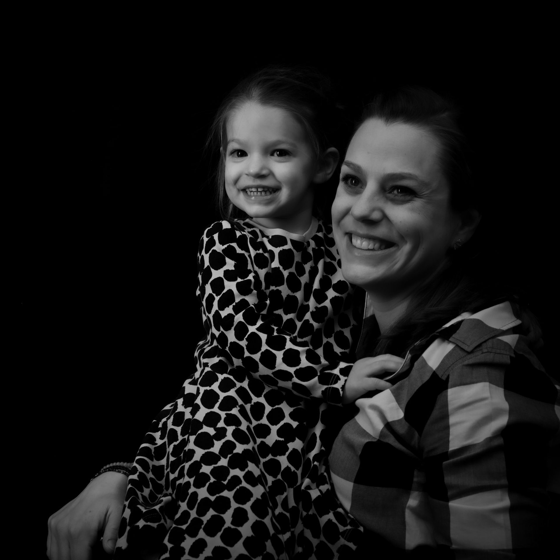 Femme et enfant souriantes et enlacées. La femme porte une chemise à carreaux. L'enfant porte une robe à pois. Fond noir.